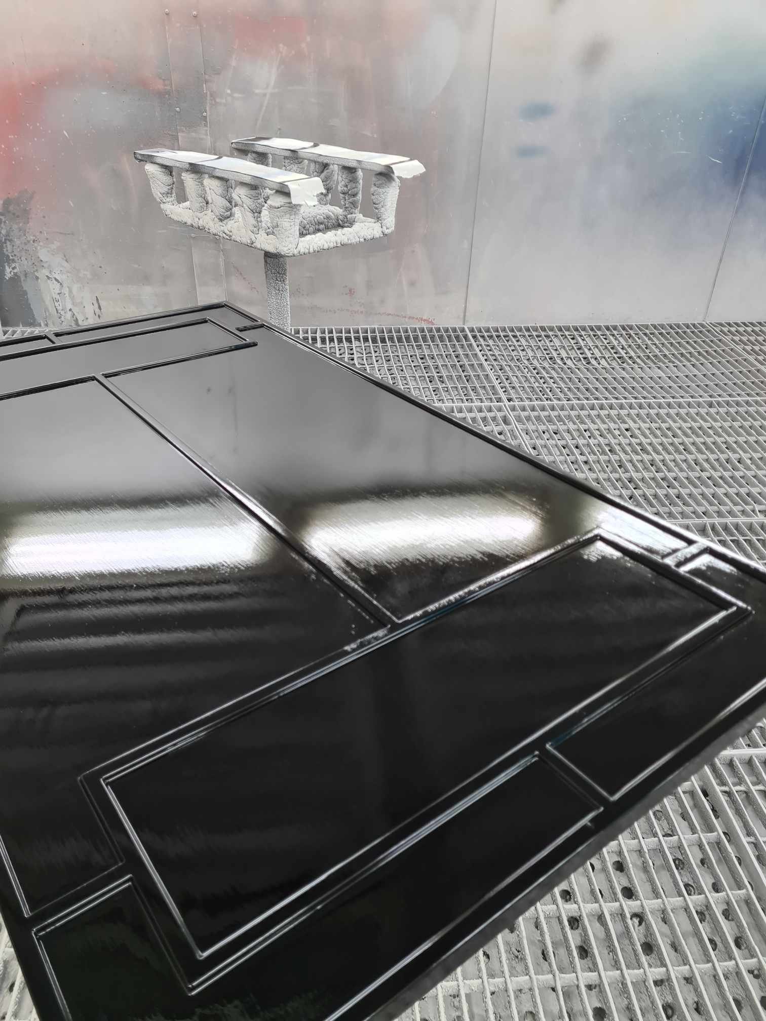 A black, glossy, freshly spray-painted object with detailed edges is in a spray booth, with a white, frosty paint sprayer in the background.