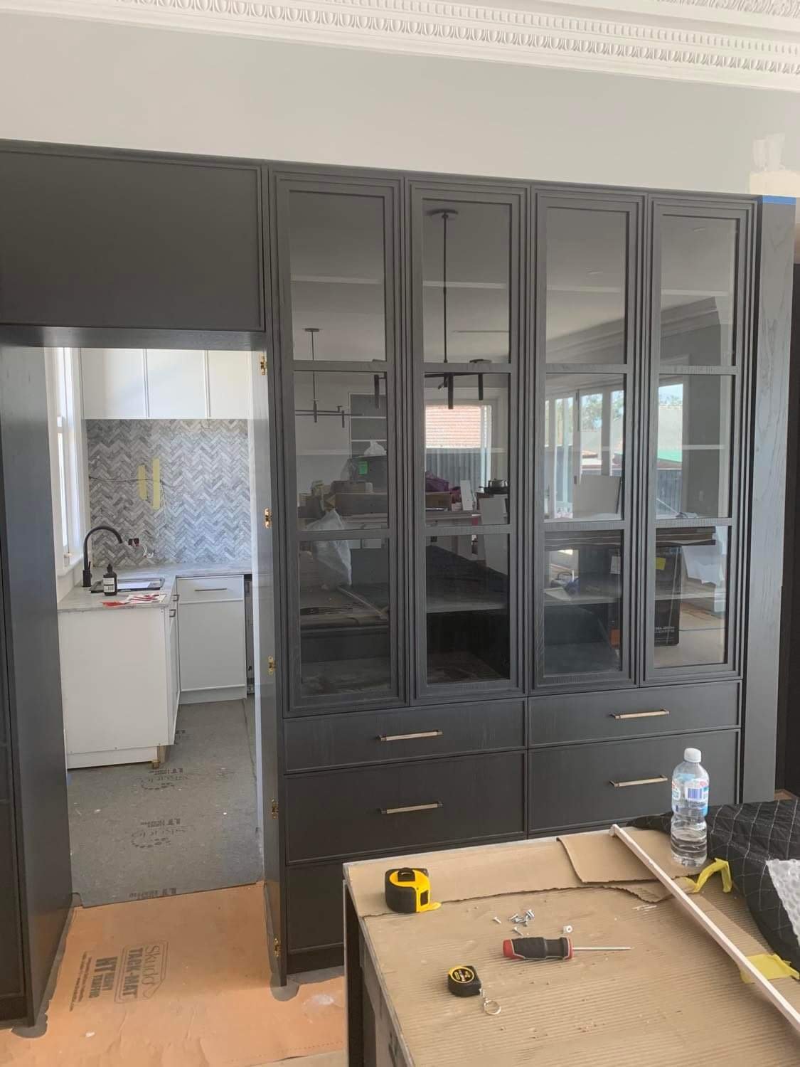 A black dining cabinet with glass doors and drawers is being assembled in a room with ongoing home renovation, with tools, a water bottle, and construction materials on a nearby table.