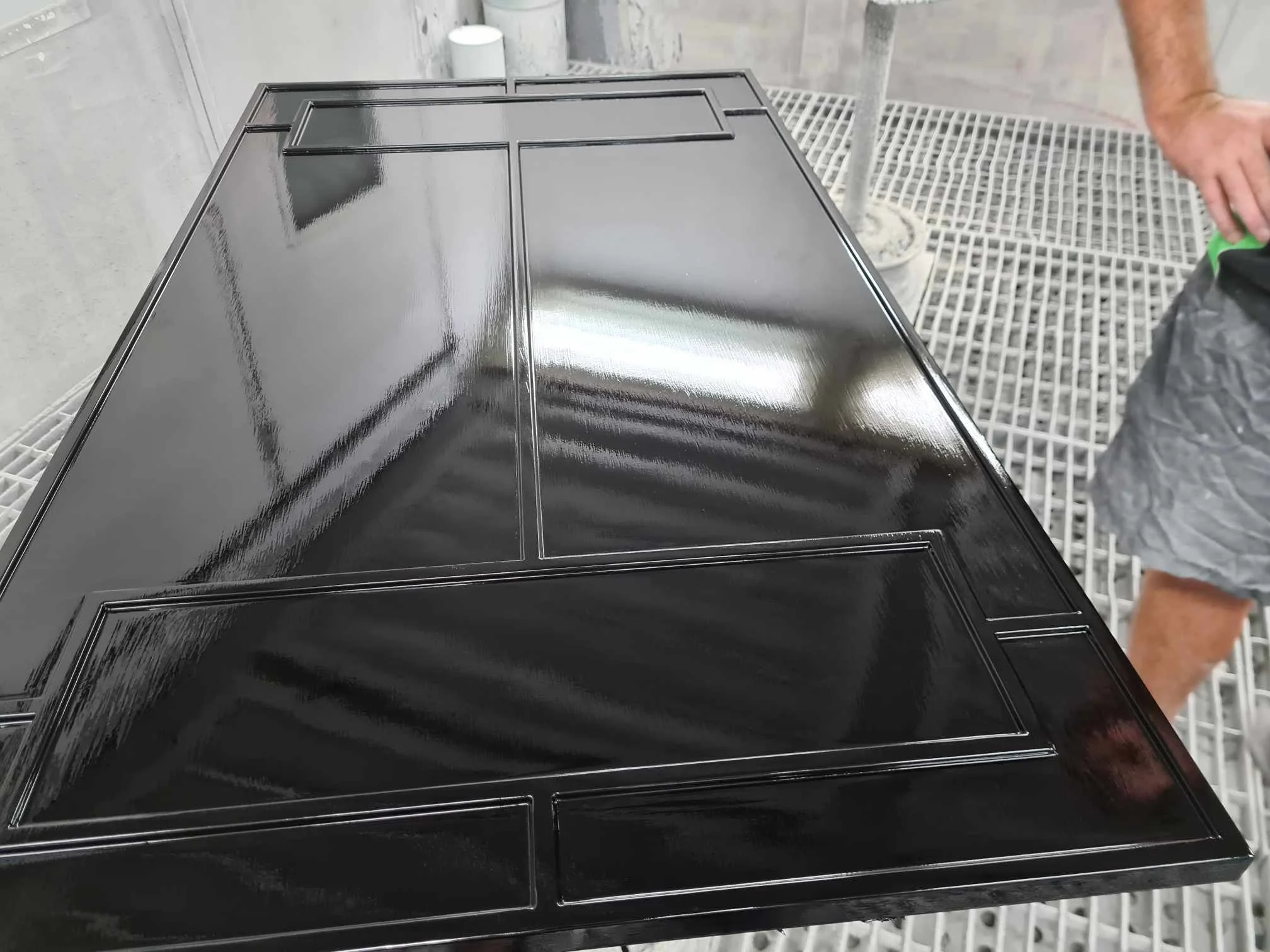 Close-up of a black, glossy, metal surface with rectangular indents, likely part of a piece of furniture or equipment, with part of a person's arm visible on the right side.
