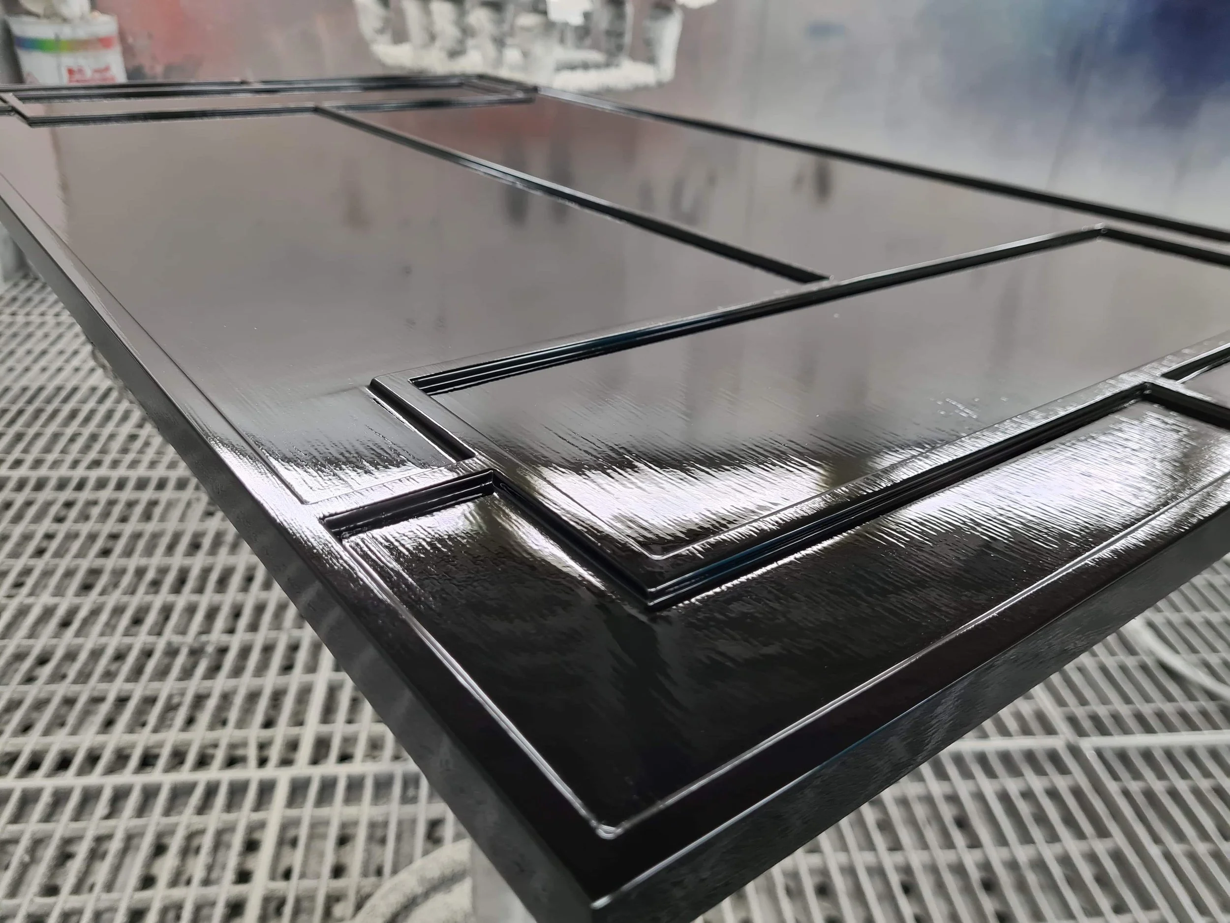 Close-up of a glossy black painted wooden table with a paneled design, placed on a grated industrial surface in a workshop or painting booth.