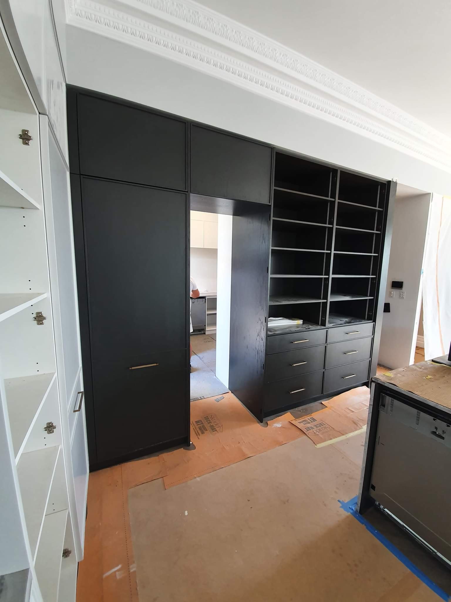 Black built-in cabinets and shelves in a room under construction, with partly visible kitchen area in the background.