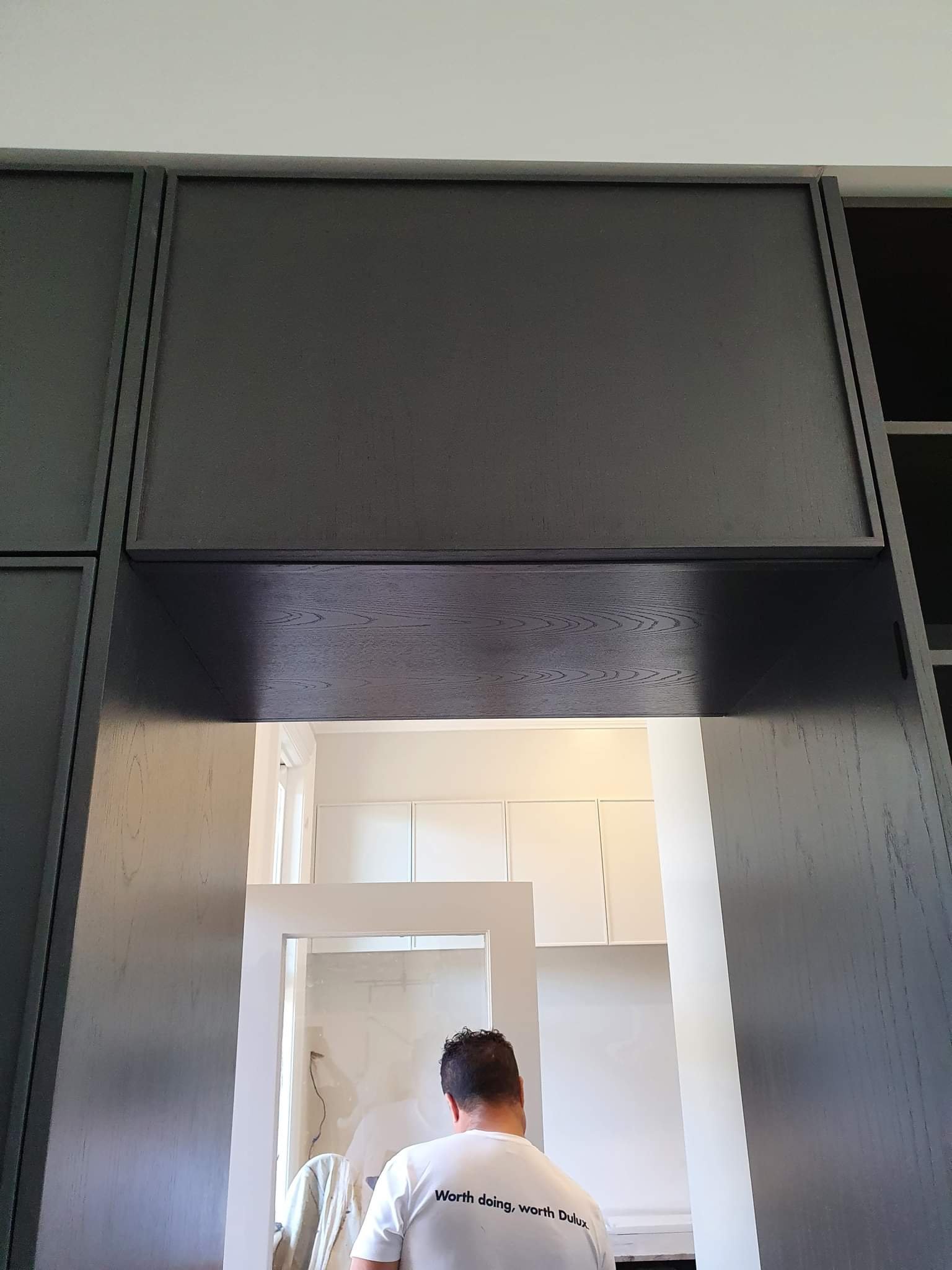 Person with short dark hair wearing a white t-shirt with the text 'Worth doing, worth Dulux' on the back, seen from behind, inside a modern interior space with a white wall and cabinet in the background.