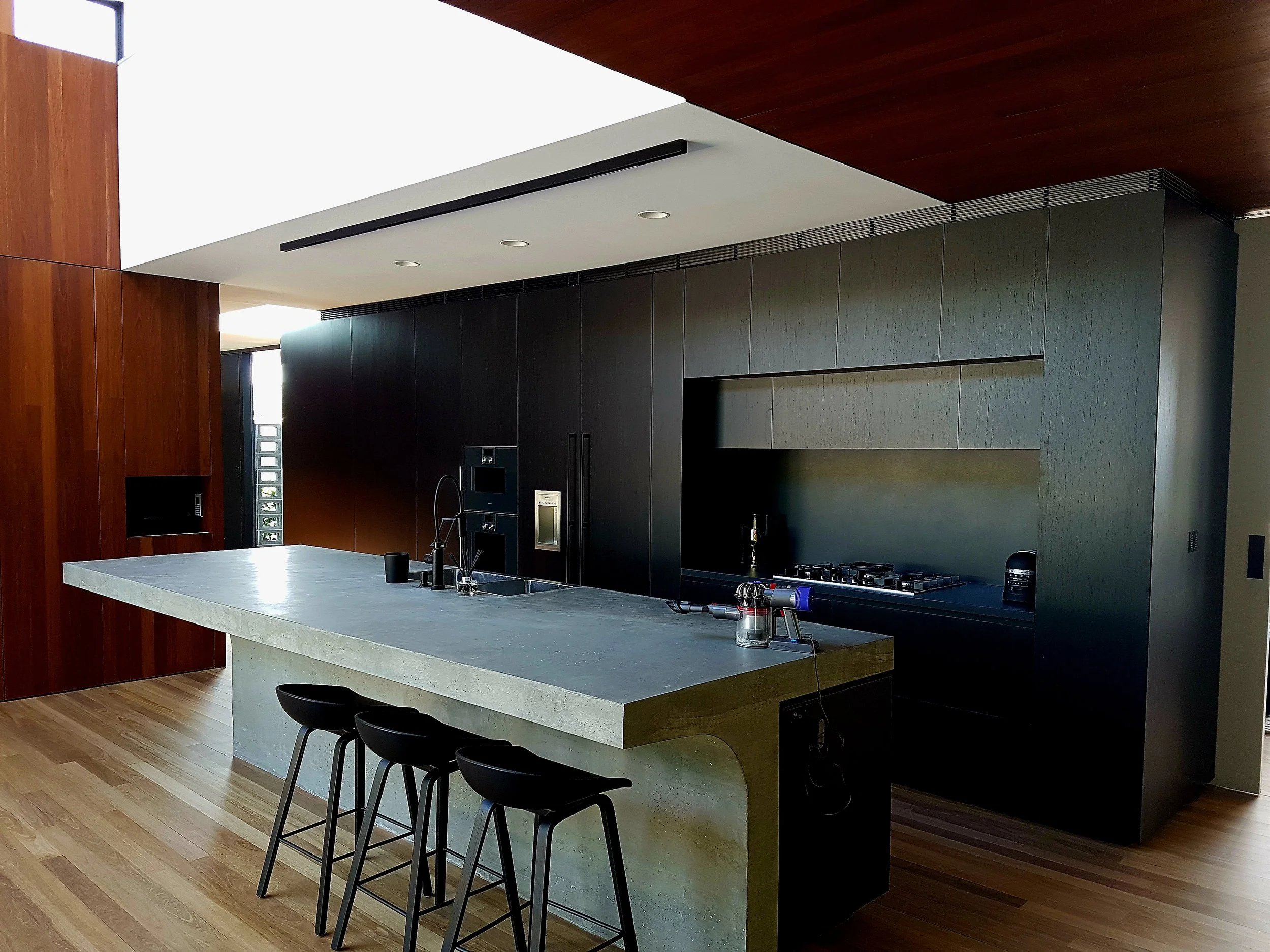 Modern kitchen with concrete island, black cabinets, built-in appliances, and wooden flooring.
