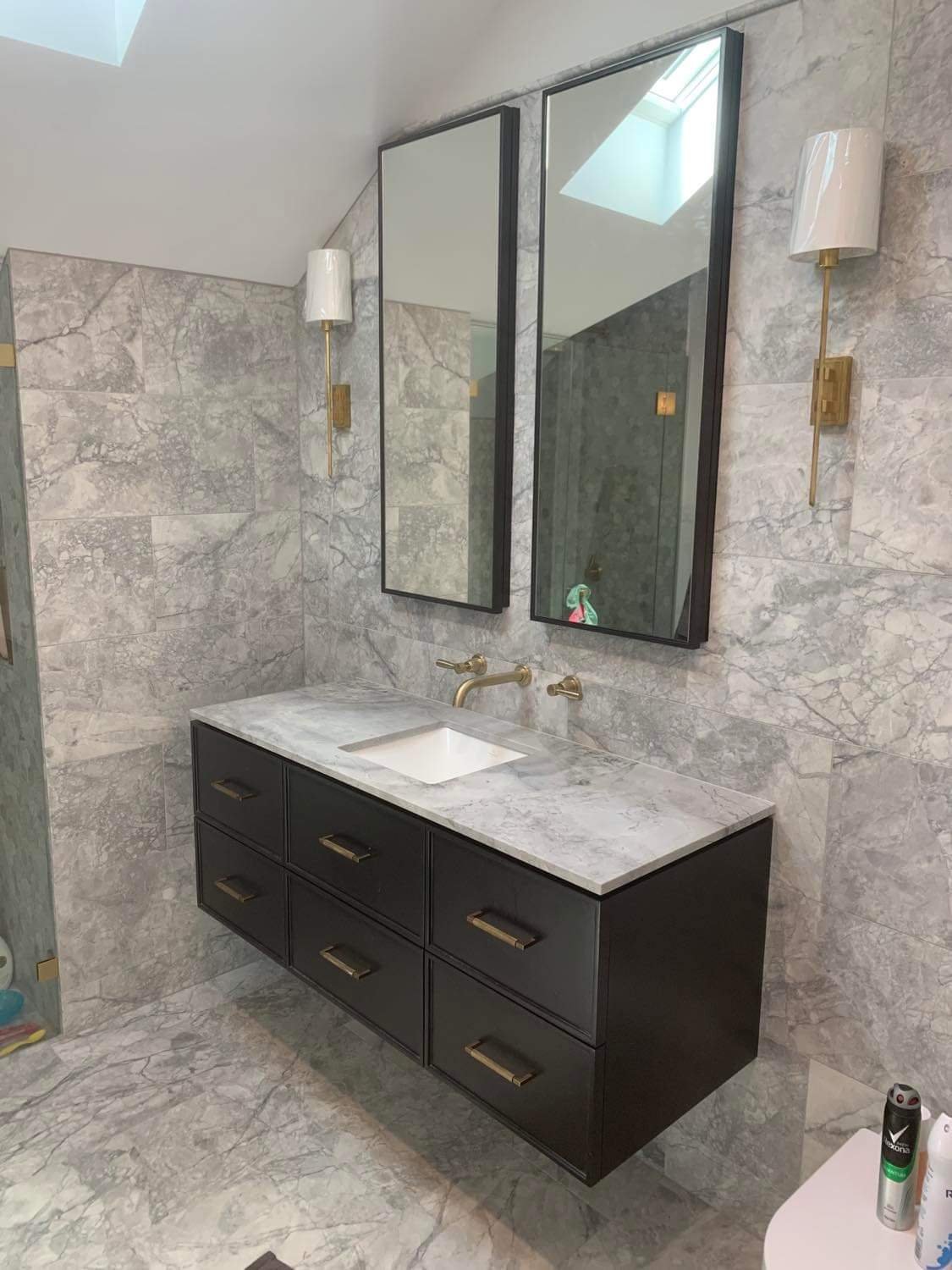 Modern bathroom vanity with black cabinet, white marble countertop, rectangular sink, and two large mirrors, with two wall-mounted gold and white sconces on a marble wall and a skylight above.
