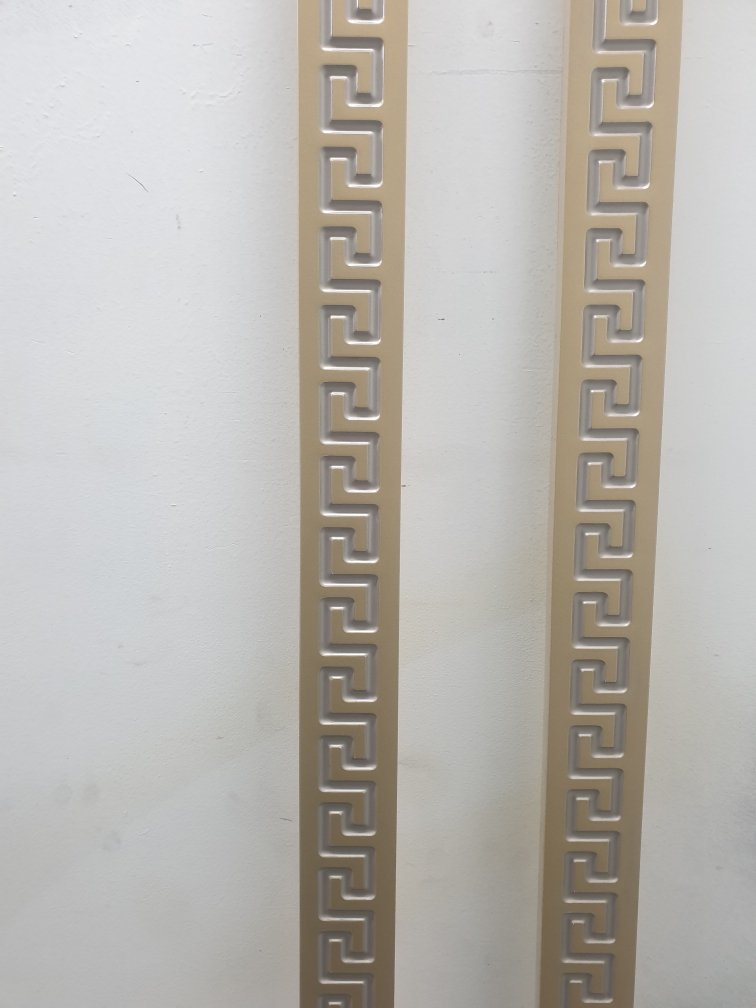 Close-up of a decorative metal panel with a Greek key pattern mounted on a white wall.