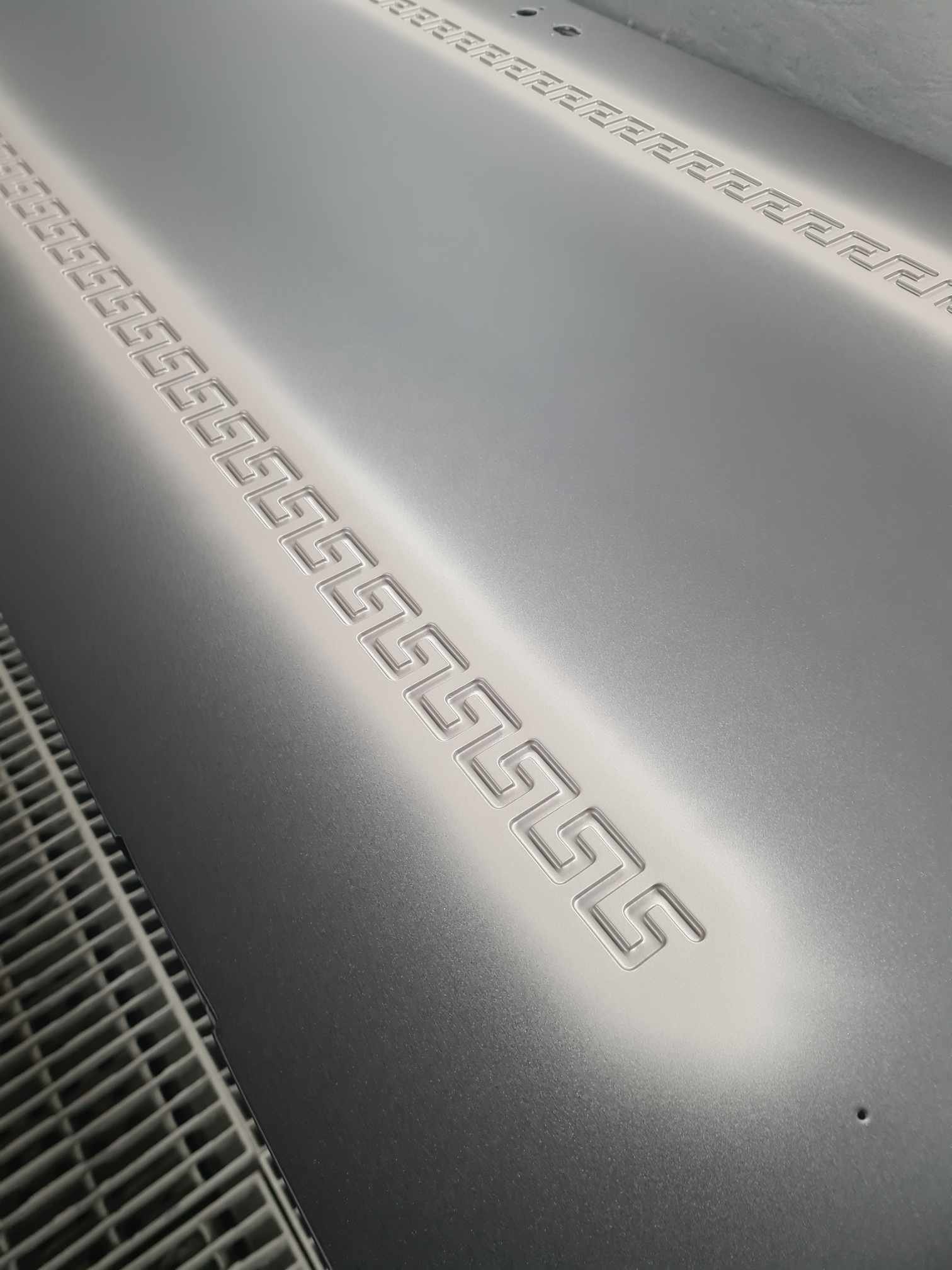 Close-up image of a silver metallic surface with an embossed geometric pattern, likely a part of a modern appliance or electronic device.