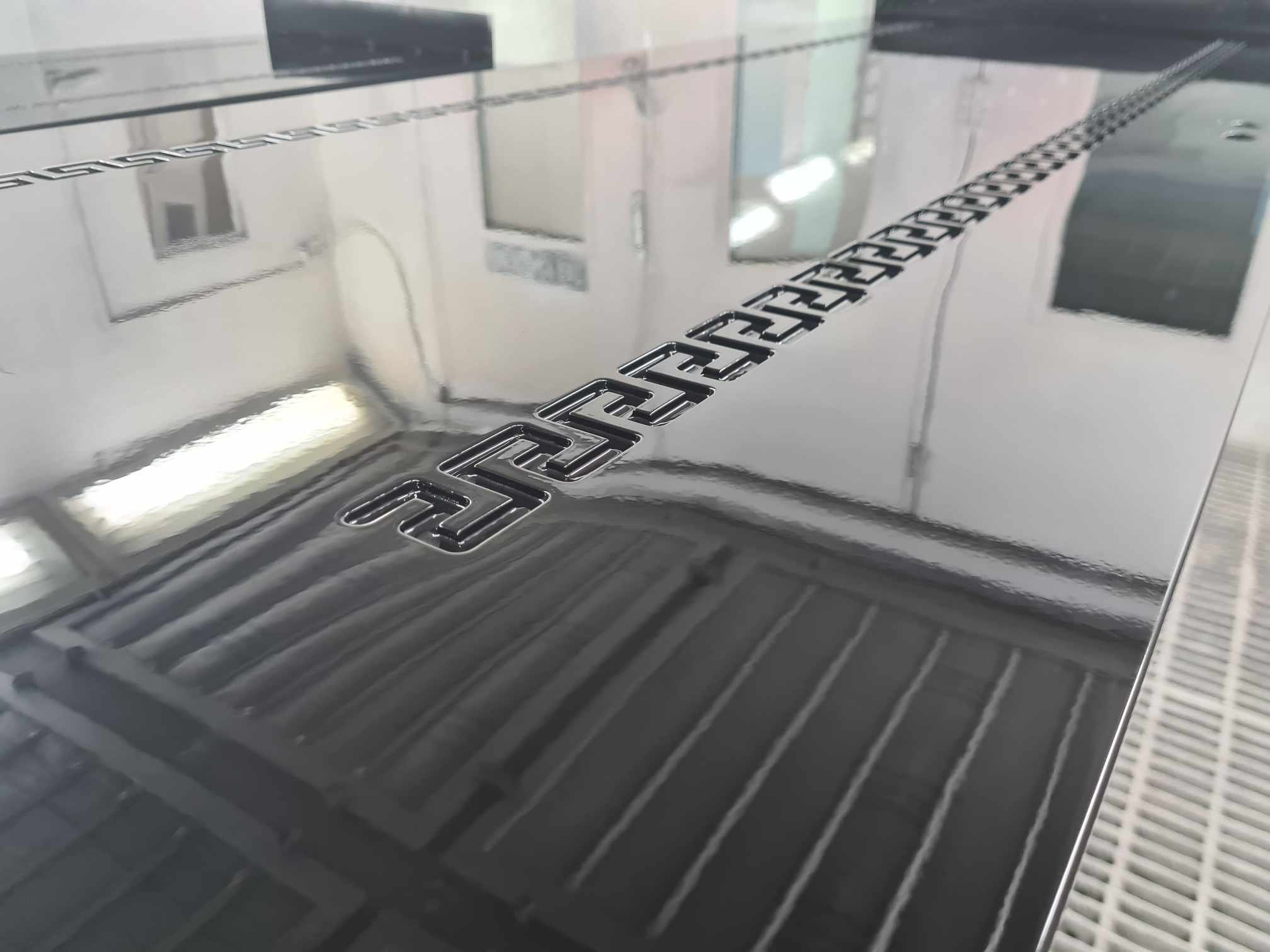 Close-up of a metallic surface with an embossed logo that says 'G Class' or similar, reflecting the surrounding room with ceilings, lights, and walls.