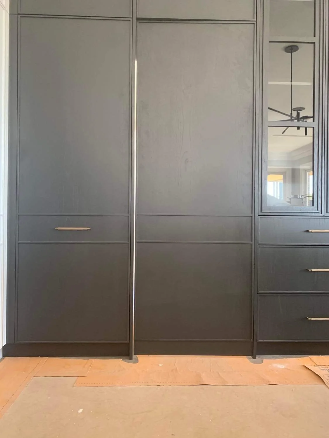 Modern black cabinetry with metallic handles in a kitchen, with a mirror and a ceiling fan visible in its reflection.