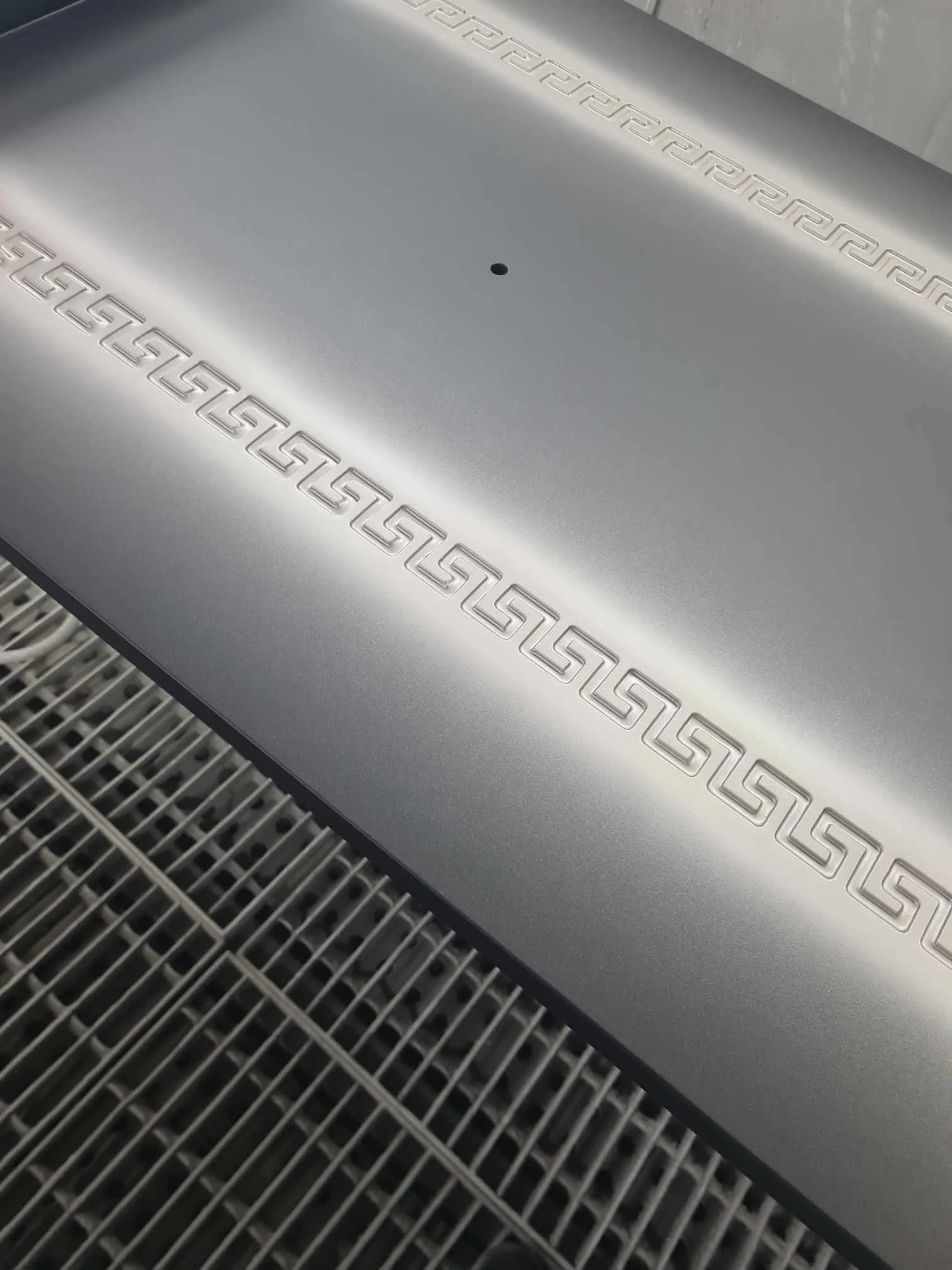 Close-up of a metallic grill with Greek key pattern embossed on a silver surface, with a small black hole in the center.