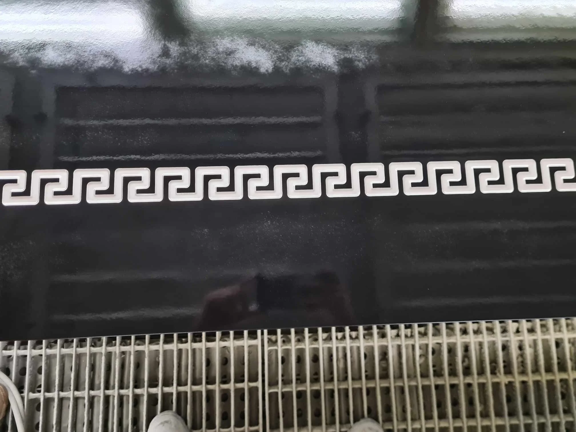 Close-up of a black surface with a white decorative border featuring a Greek key pattern, with part of gray footwear visible at the bottom.