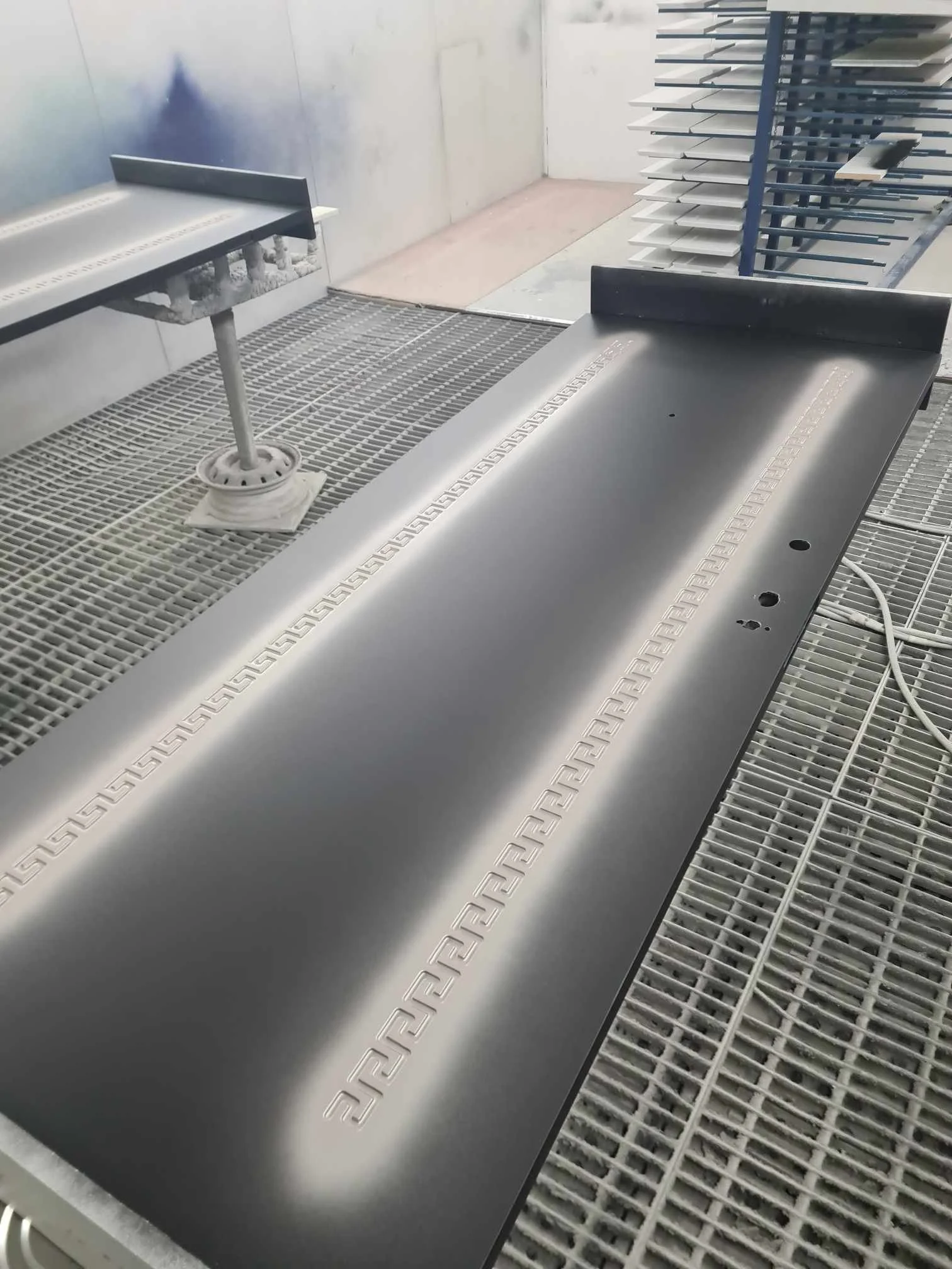 A metallic, matte black baseball bat with Greek key border design along the top edge resting on a white ventilated work surface in a manufacturing or coating facility.