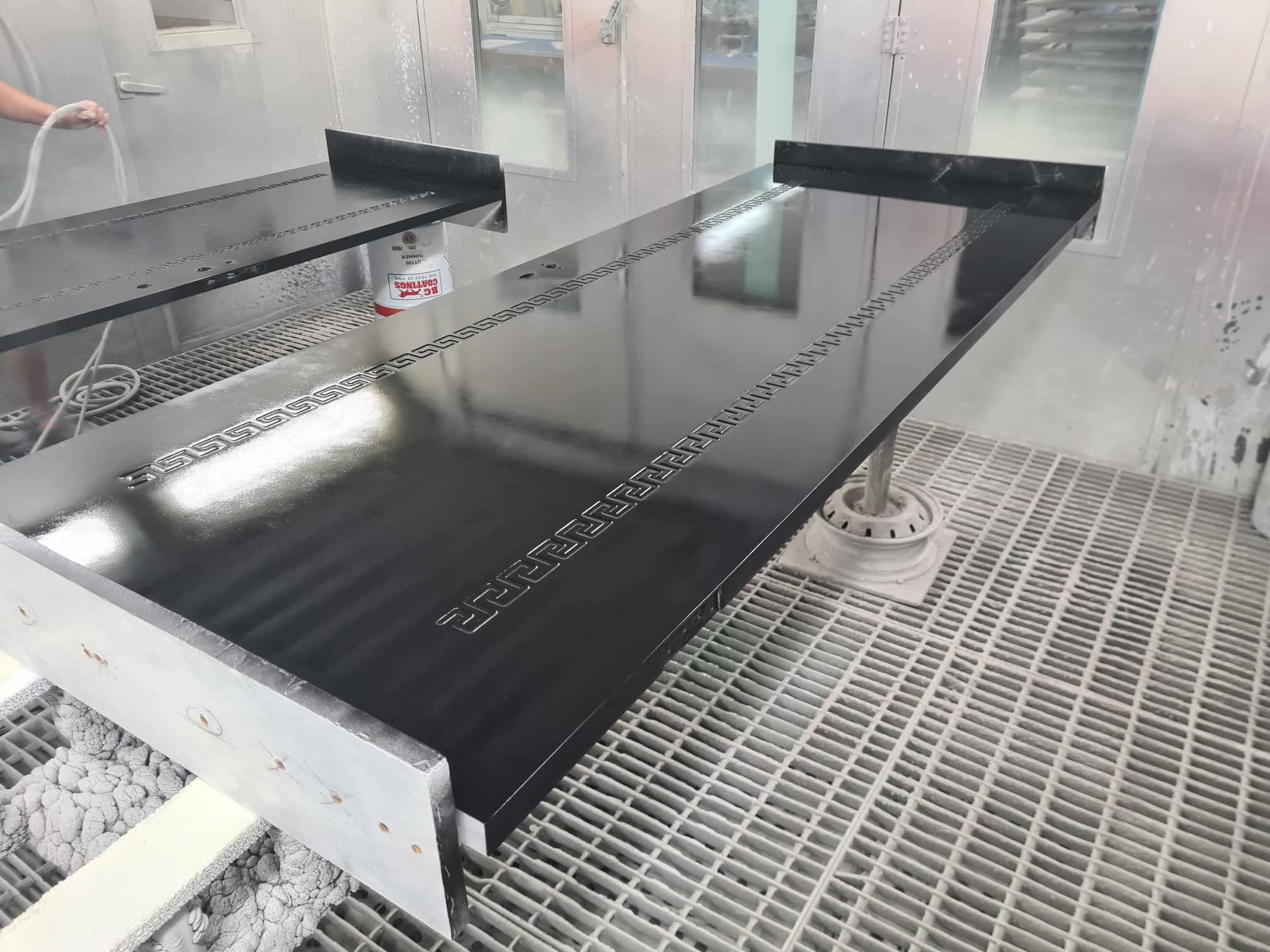 A black metal countertop with an embossed Greek key pattern along the edges is being painted or coated in a spray booth with a person holding a spray gun in the background. The surface is glossy and reflective.