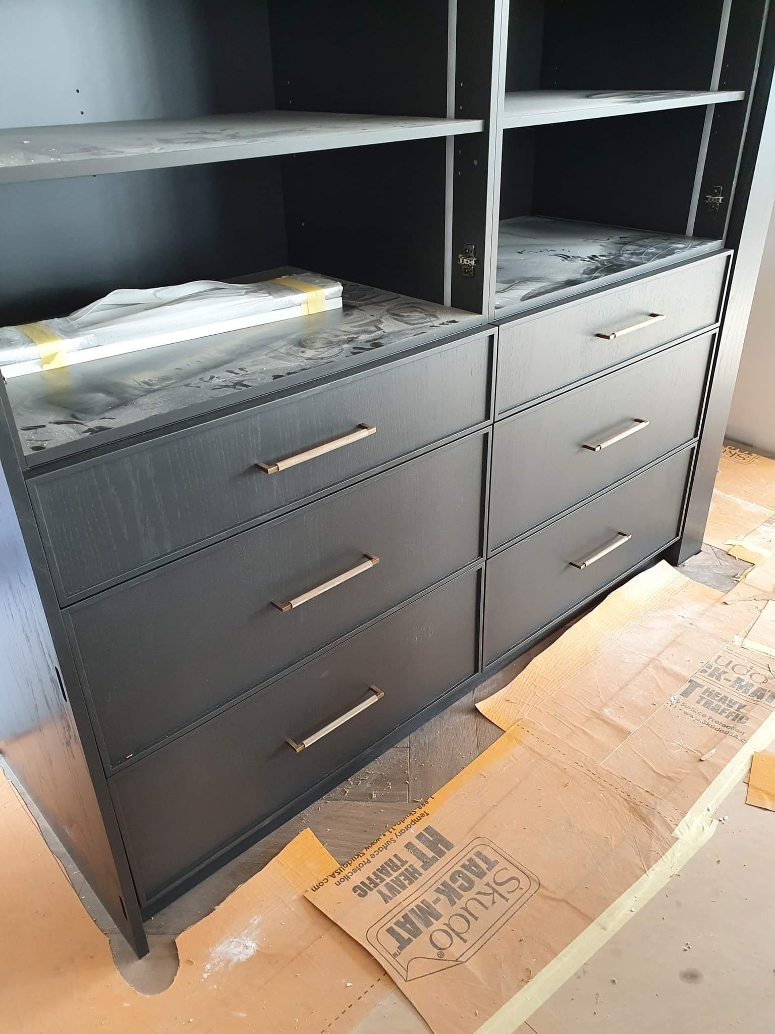 A black dresser with multiple drawers and open shelves, partially assembled, on a floor covered with cardboard meant to protect the surface during construction.