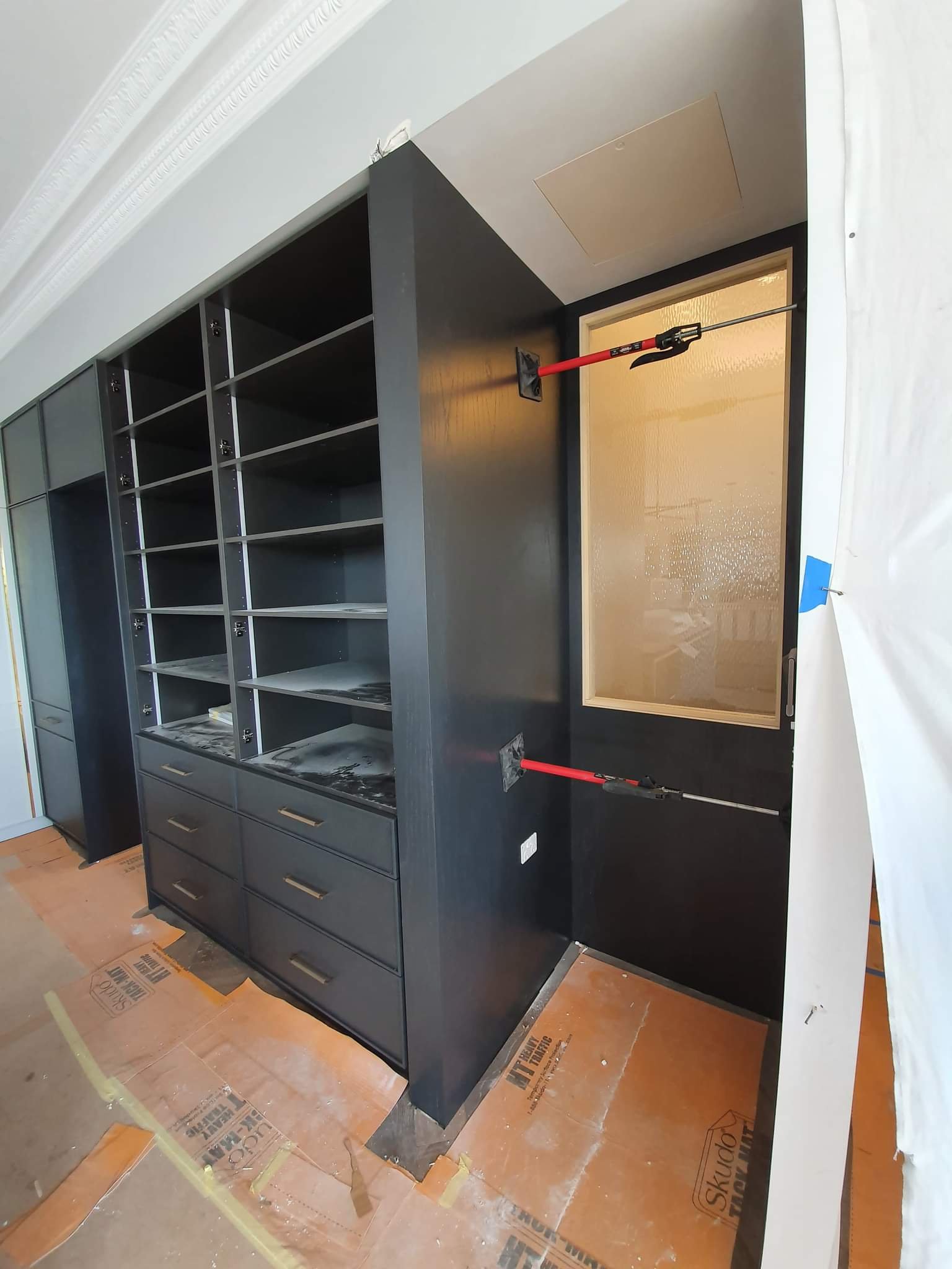A room under construction with black shelving units, a window, and two red support poles attached to the wall around the window.