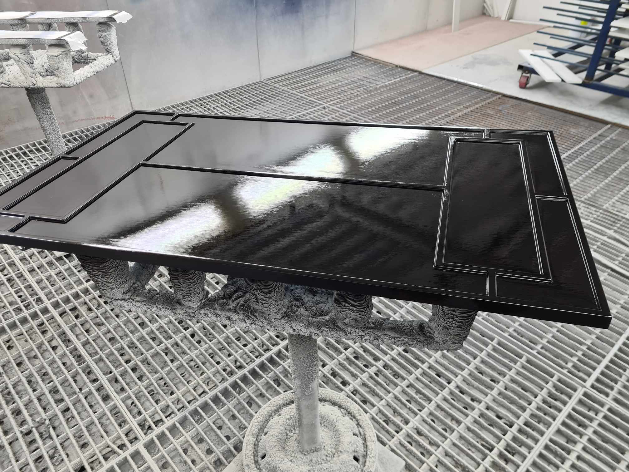 A black storage or display case with a glossy finish, placed on a painted metal table with a grid surface, in a workshop or paint booth.