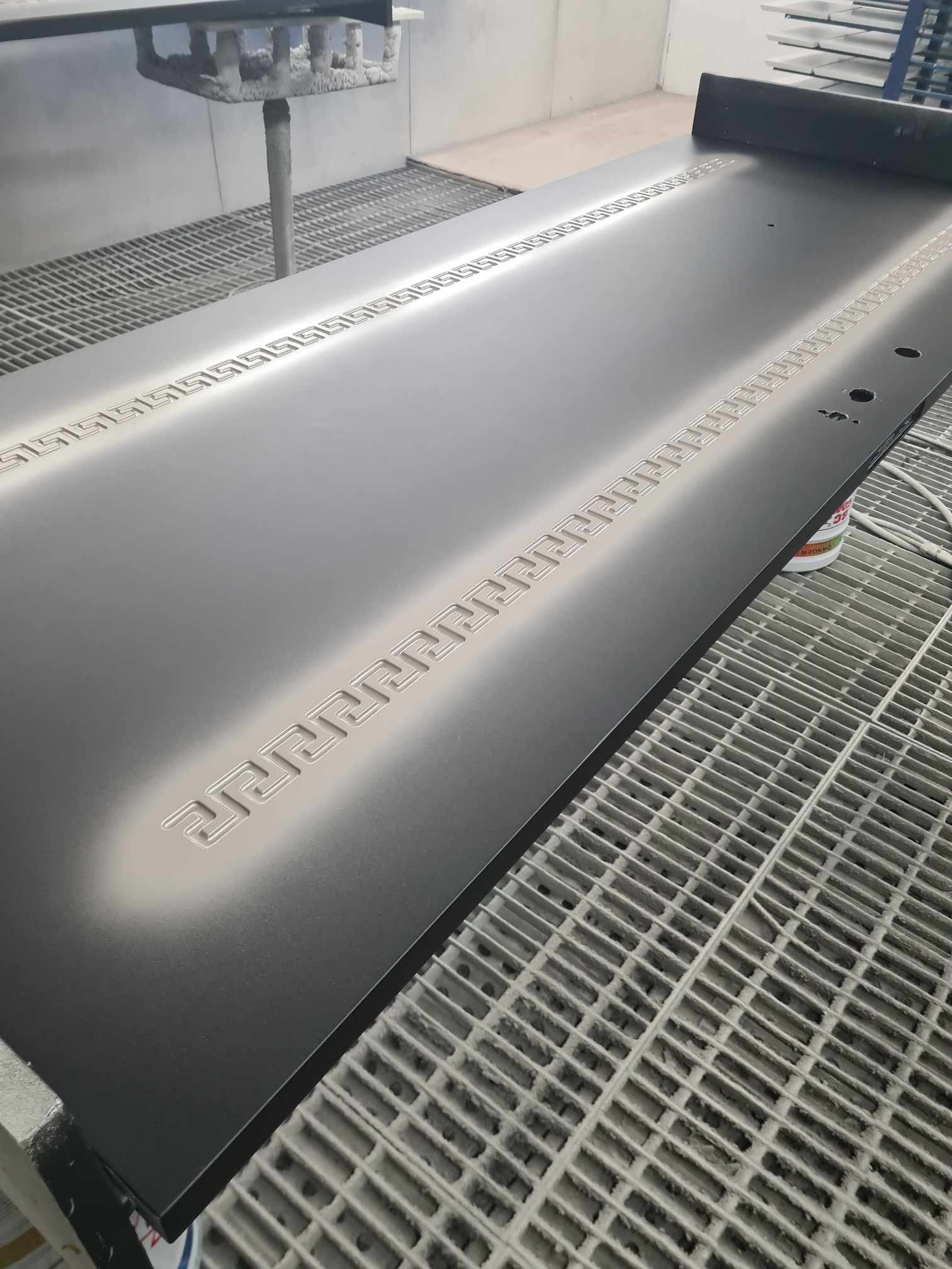 An unpainted metallic surfboard with a Greek key pattern along the edges, placed on a workbench with painting supplies in the background.