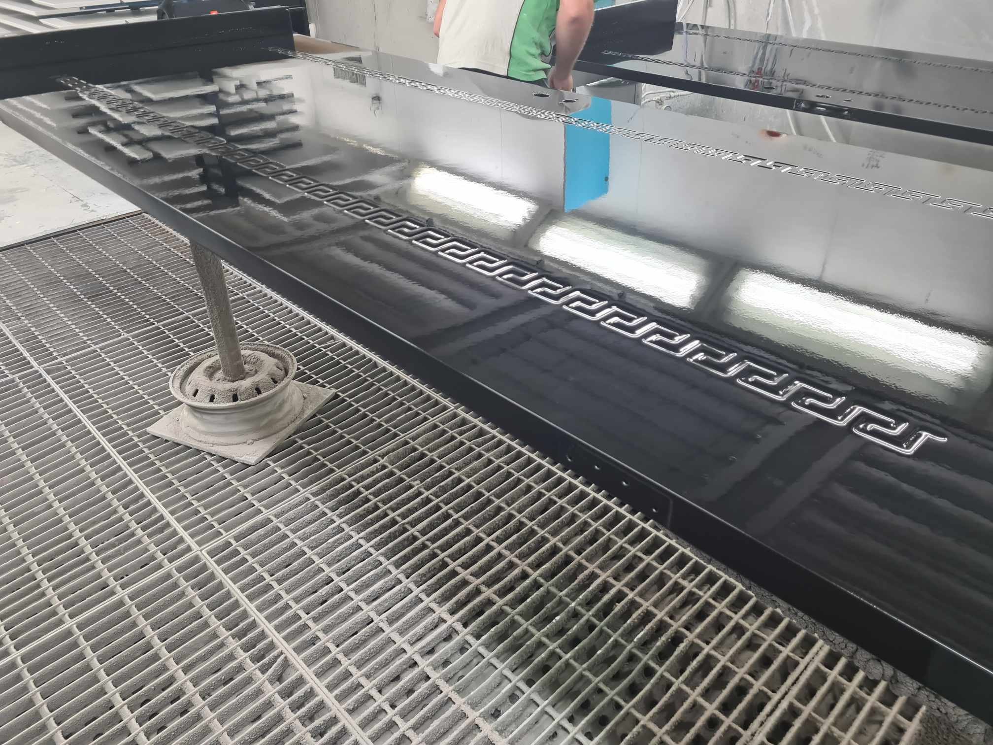 A large, shiny, black metal mold with a Greek key pattern on top, positioned above a metal grate in an industrial setting.