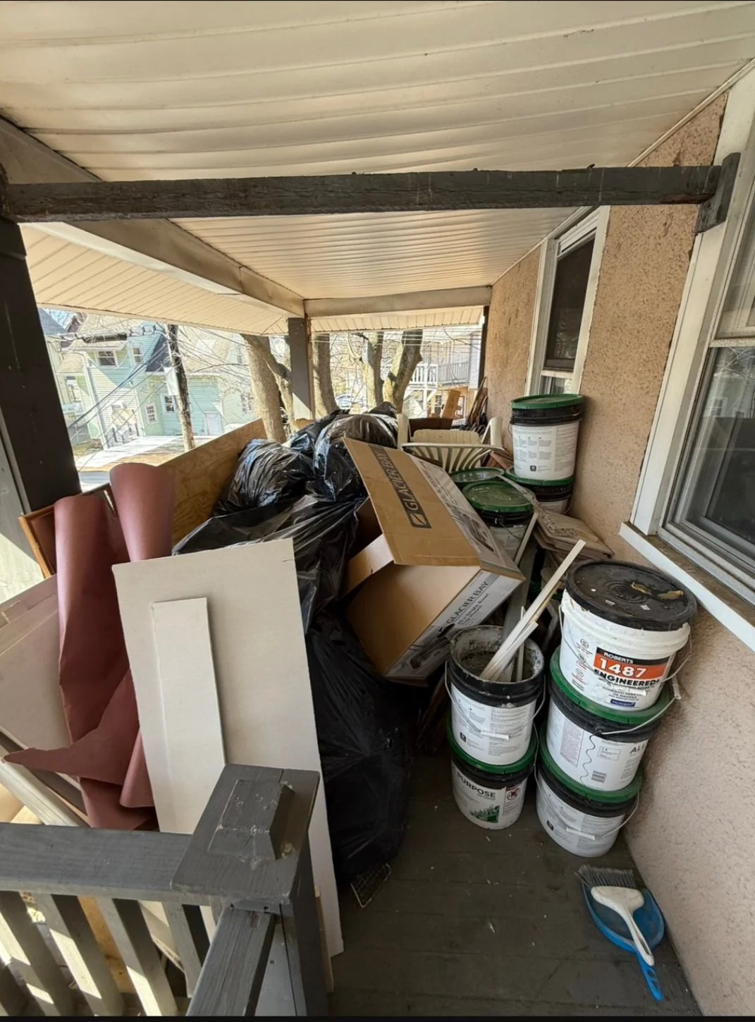 A cluttered balcony with buckets of paint, cardboard boxes, black trash bags, and various household items.