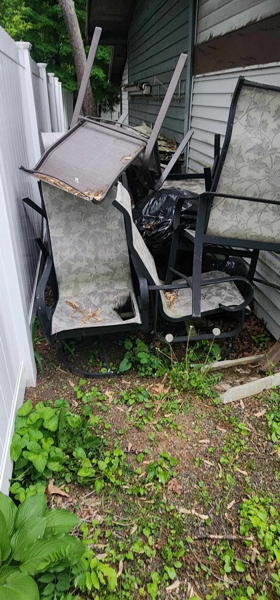 A pile of overturned outdoor chairs, including glass-top tables and cushioned seats, stacked against the side of a house with disturbed vegetation on the ground.