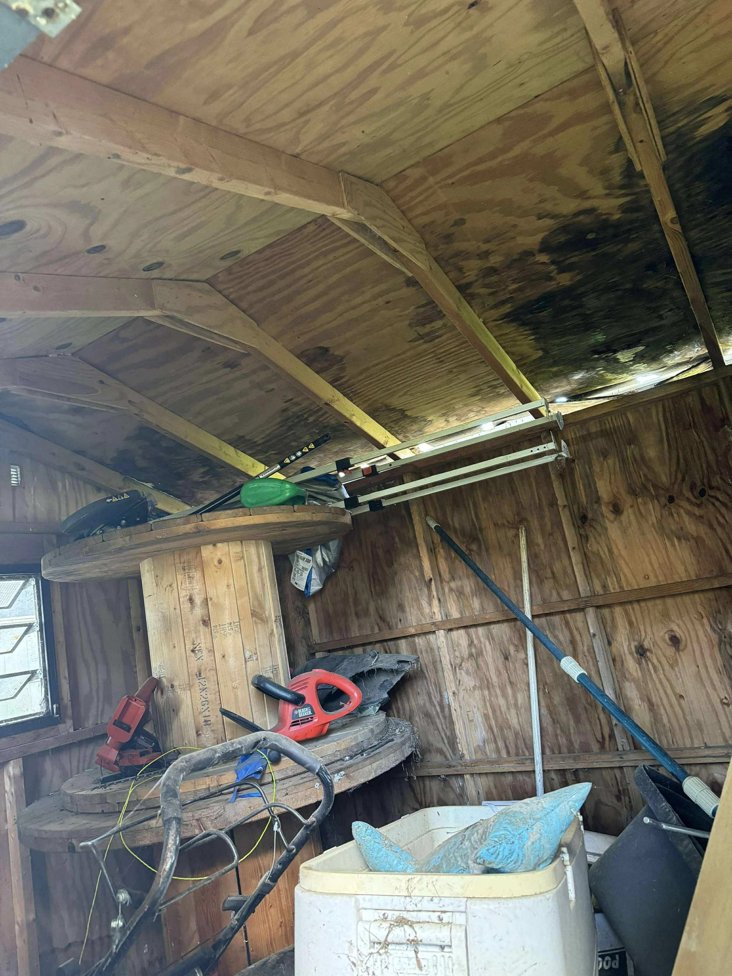 Interior of a garage with wooden walls and ceiling, cluttered with tools, a chainsaw, a broom, a shovel, and miscellaneous items.