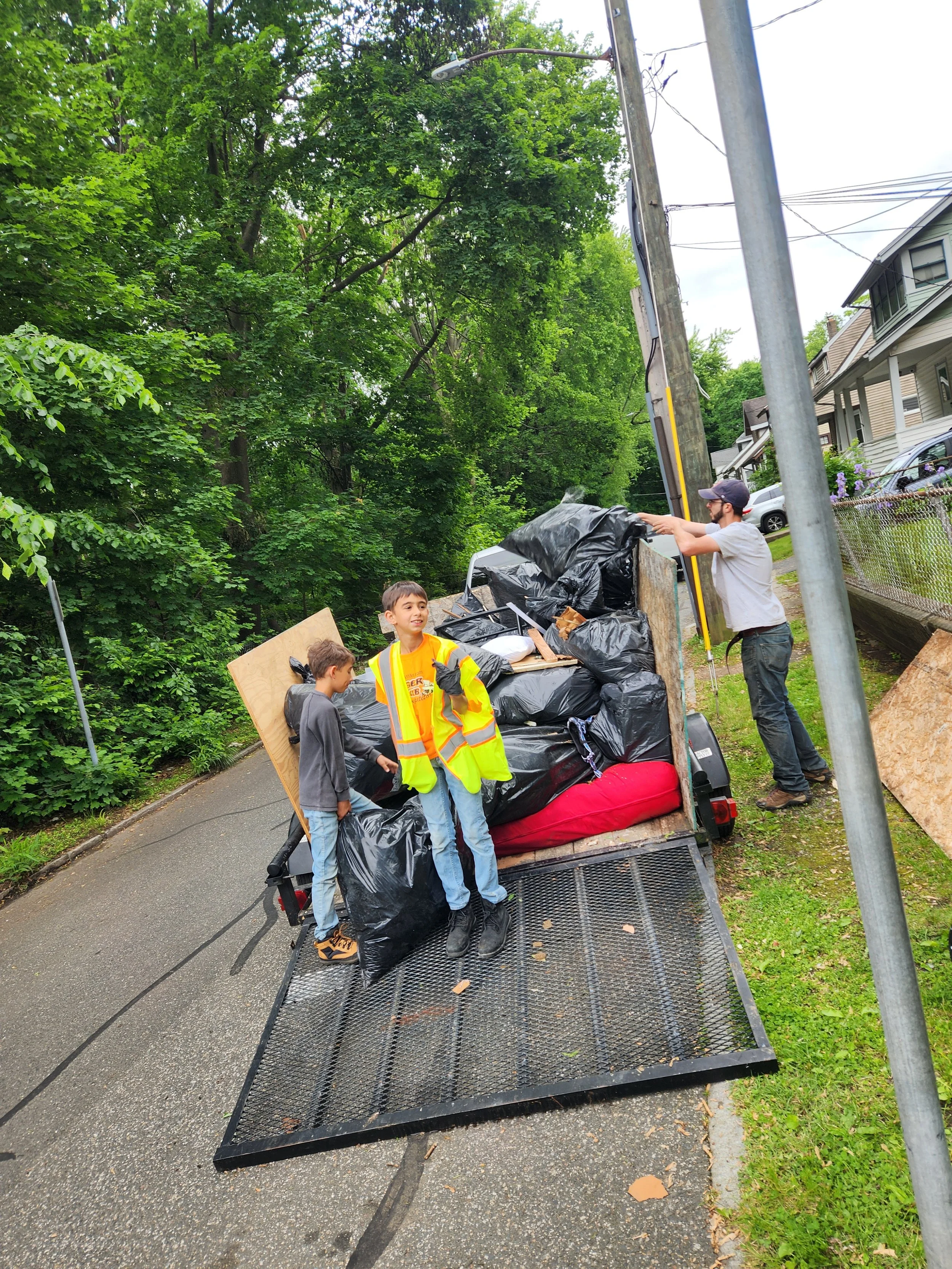 Two boys and one man standing on a trailer filled with black trash bags, plywood, and other debris, next to a residential street lined with trees and houses, possibly during a cleanup or moving activity.