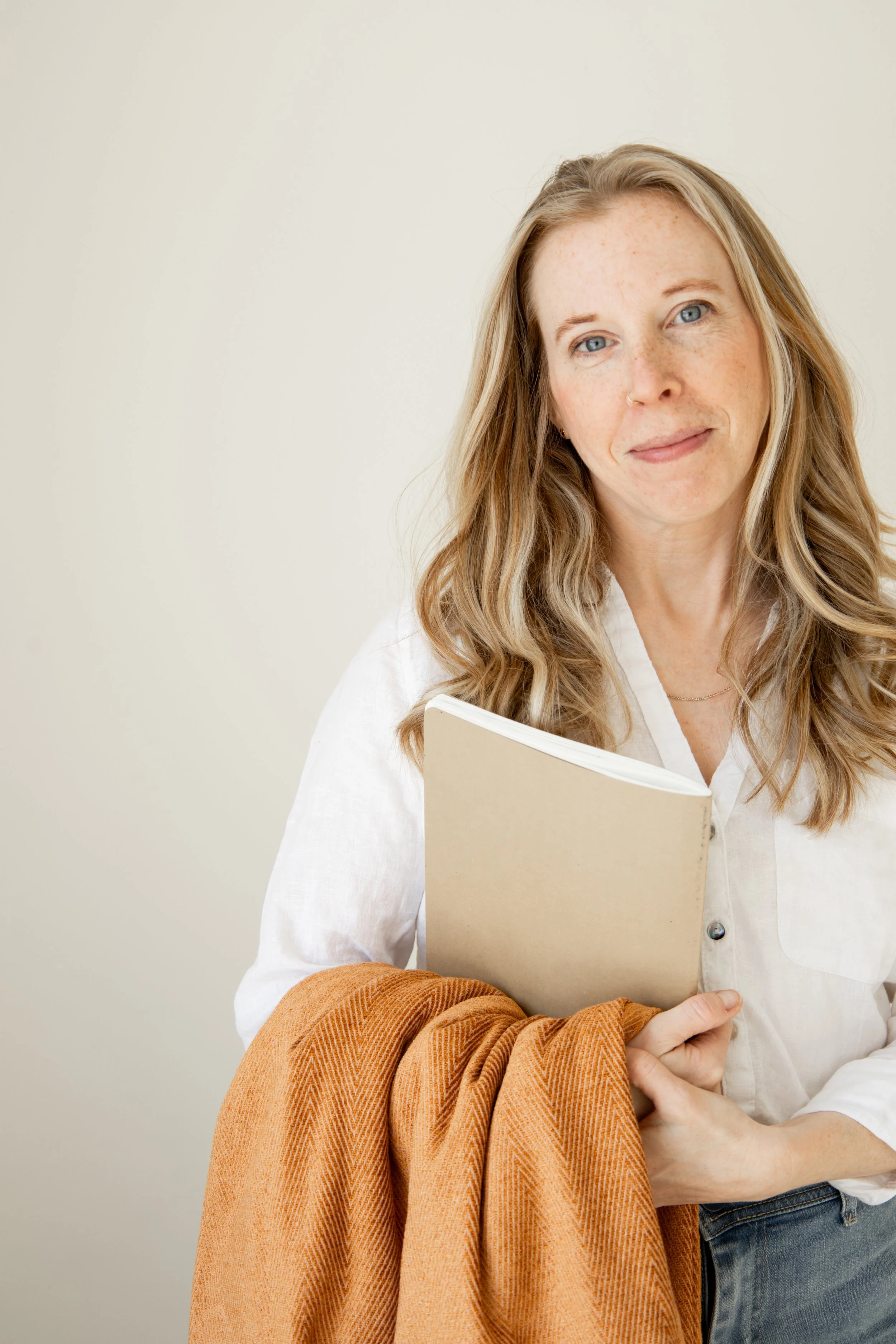A white woman with long blonde hair and blue eyes holding a beige notebook and a brown blanket over her arm, standing against a plain light-colored wall.