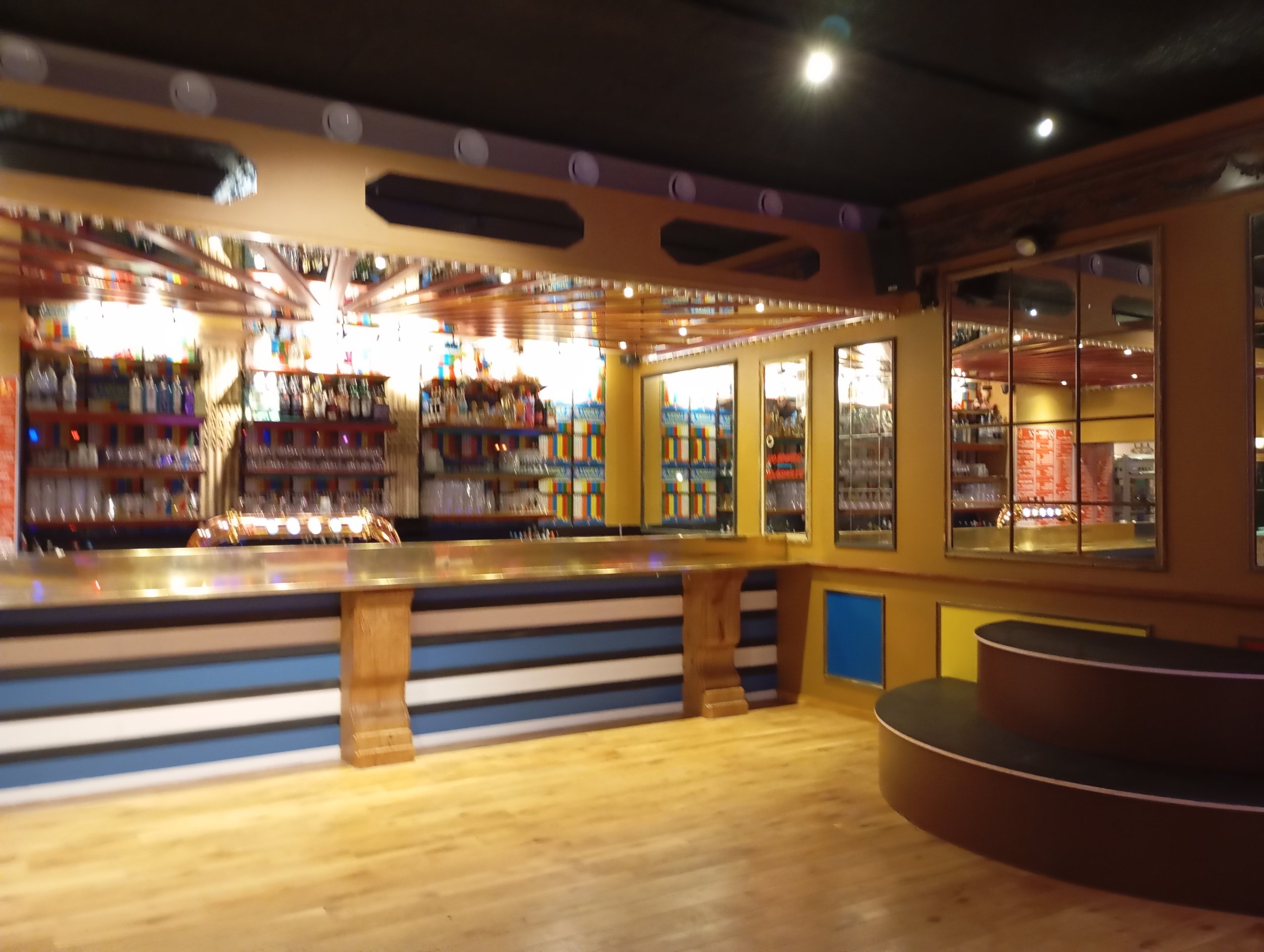 Intérieur du bar dansant avec un comptoir en bois, étagère à alcool et pompes à bière, remplies de livres ou d'objets colorés, murs en jaune avec des fenêtres miroir et un plafond en bois.