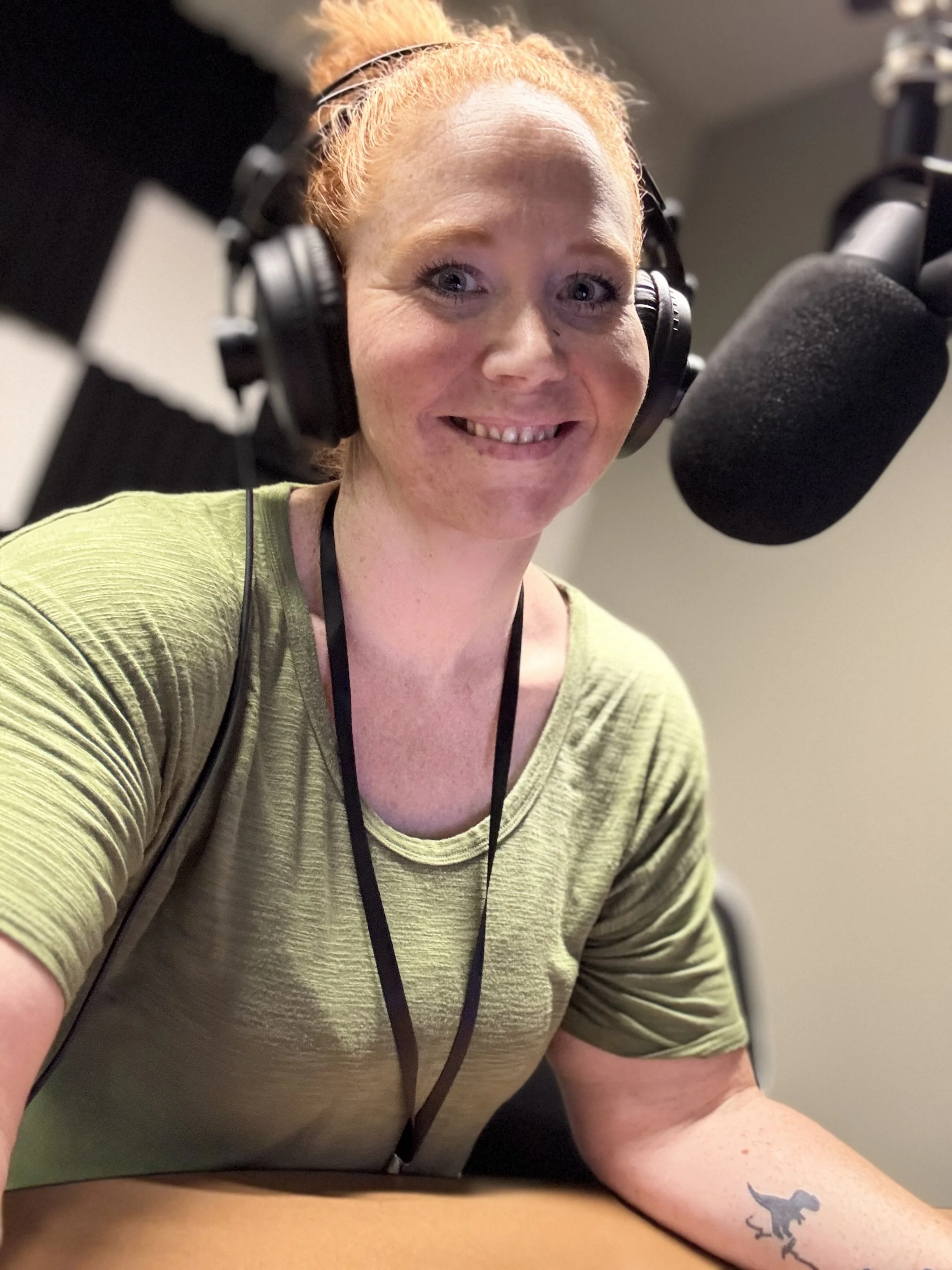 A woman with red hair and freckles, wearing a green shirt and a black lanyard, recording in a soundproof studio with a microphone and headphones.