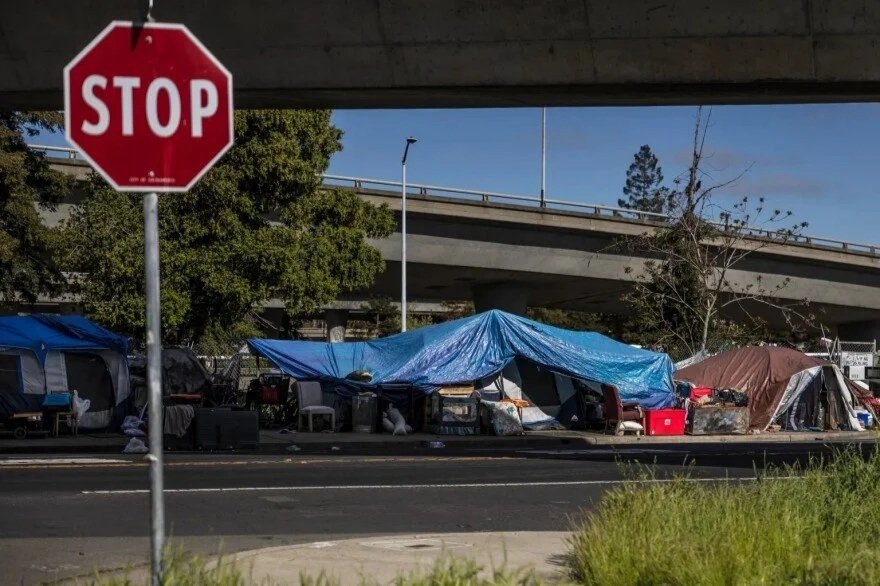 A homeless encampment under a bridge with tents covered in blue and brown tarps, and various belongings scattered around. A red stop sign is visible in the foreground.