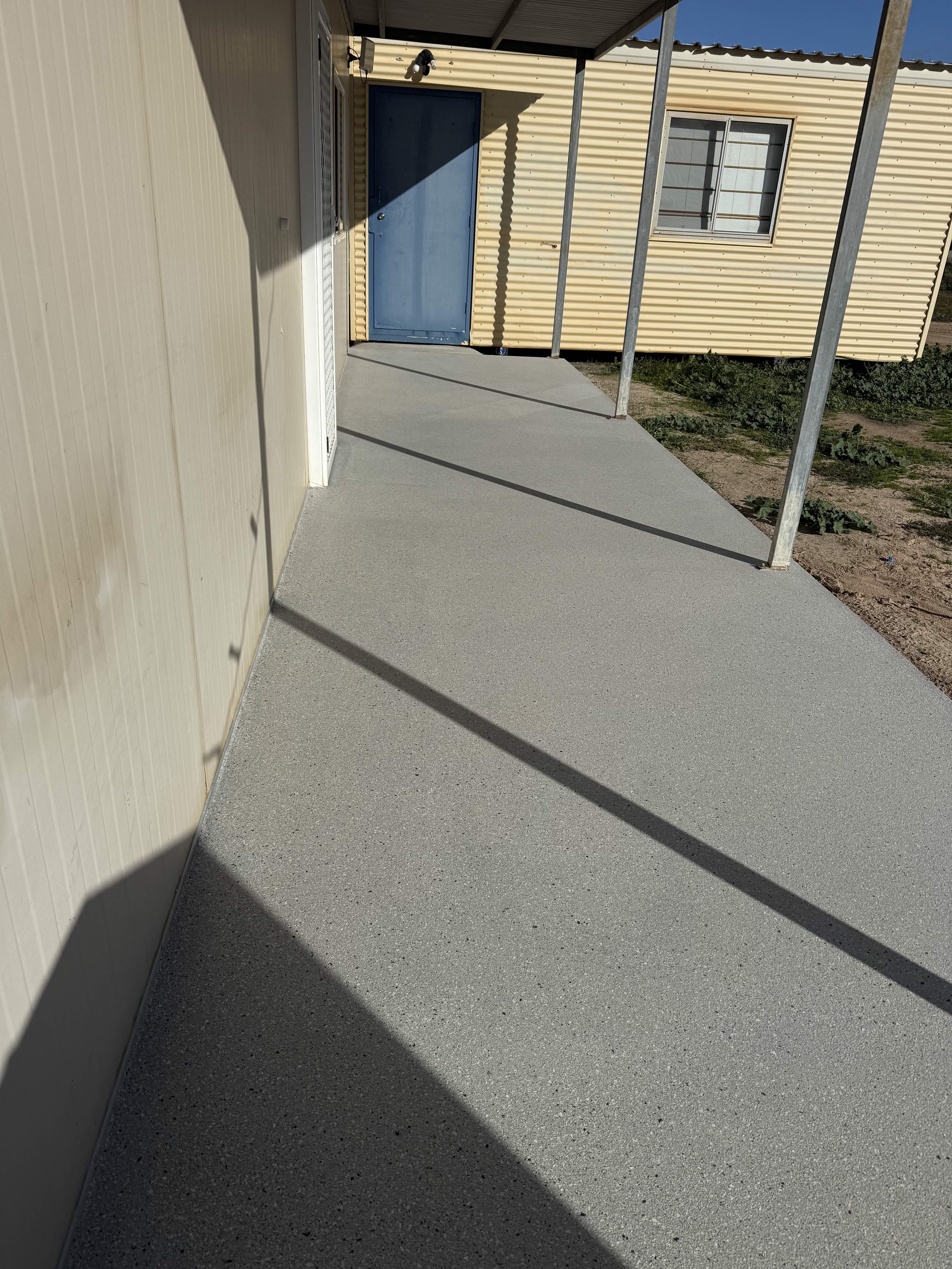 Epoxy flake walkway with fresh gray concrete leading to a blue door on a yellow metal building, with metal support beams and a window in the background.