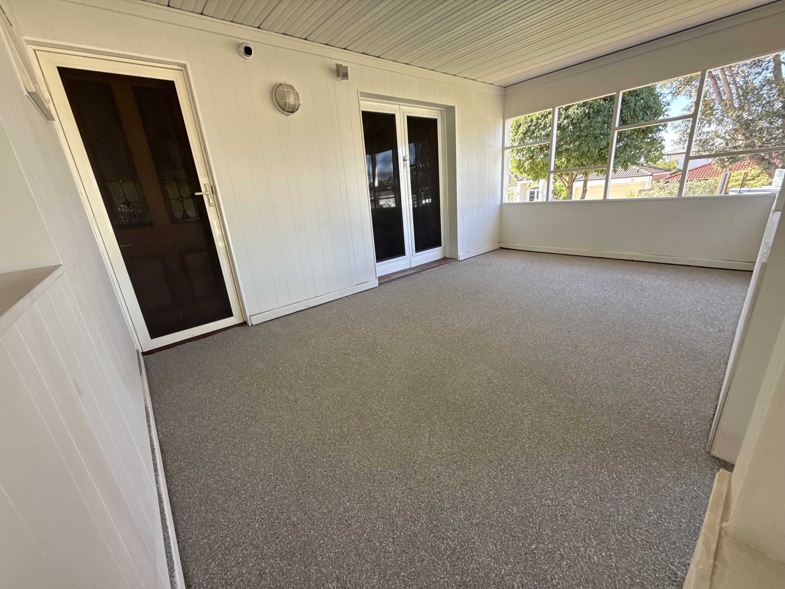 Epoxy flake balcony or sunroom with white panel walls, beige carpet, and large windows showing trees and neighboring houses.