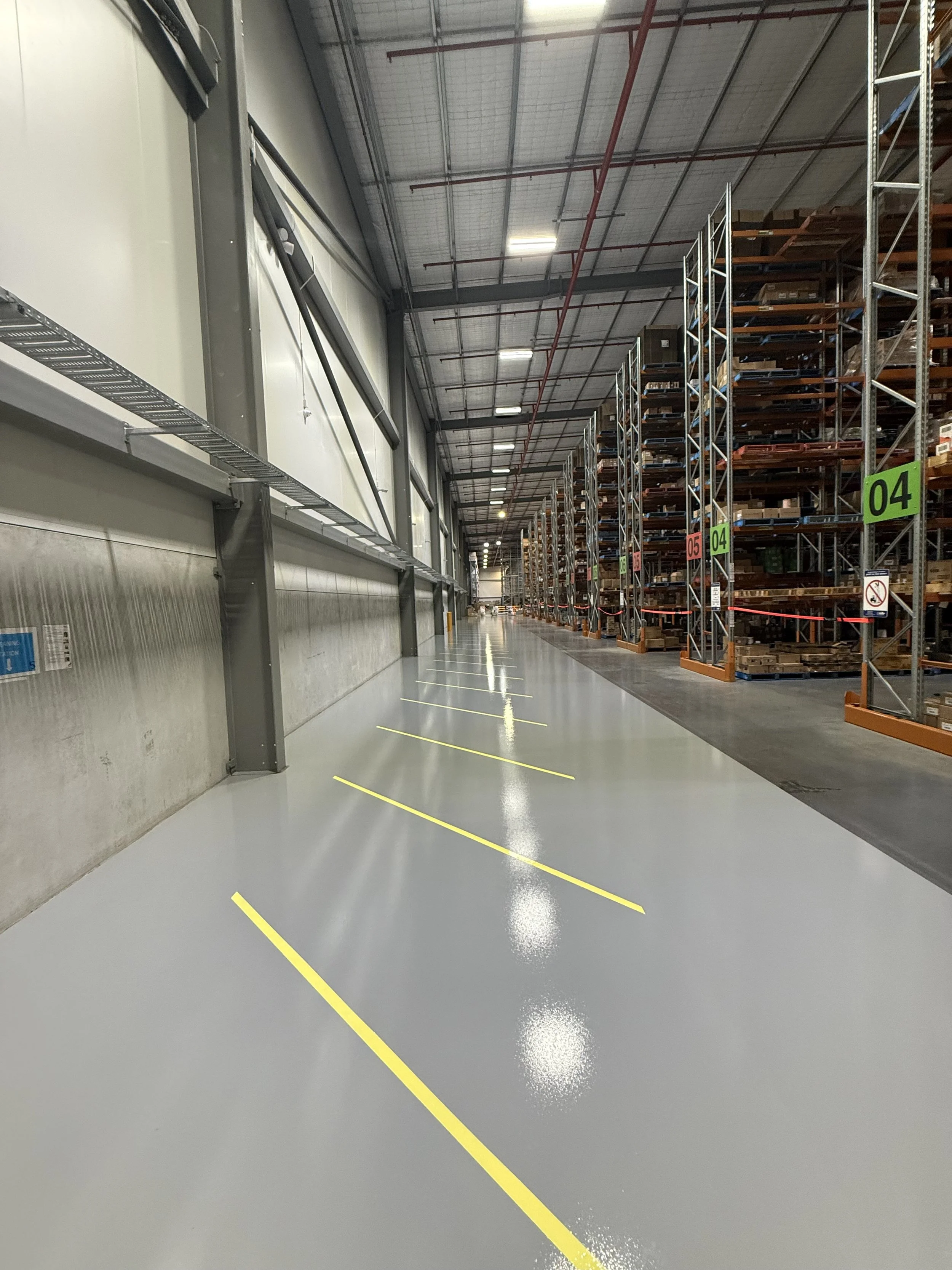 Commercial warehouse epoxy flooring installation with full concrete grinding, epoxy priming, seamless grey epoxy coating, and precision yellow line marking. This industrial warehouse floor was professionally prepared and coated to create a durable, e