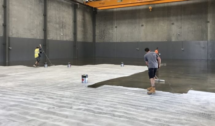 Two workers are painting epoxy onto a large concrete floor inside an industrial building with exposed concrete walls and a yellow overhead crane.