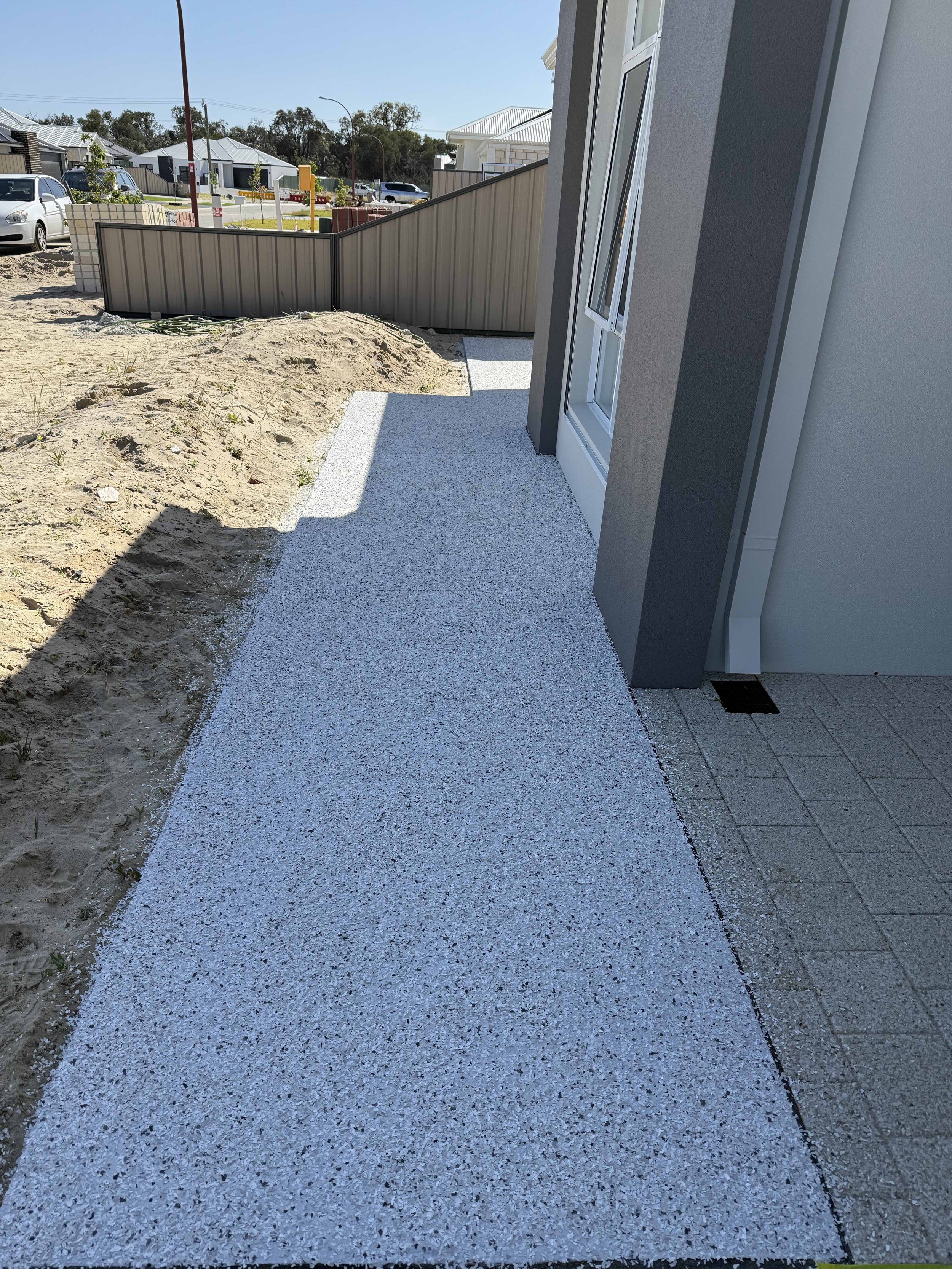 Epoxy flake sidewalk next to a house with a window, with some dirt and landscaping on the side, in a suburban neighborhood.
