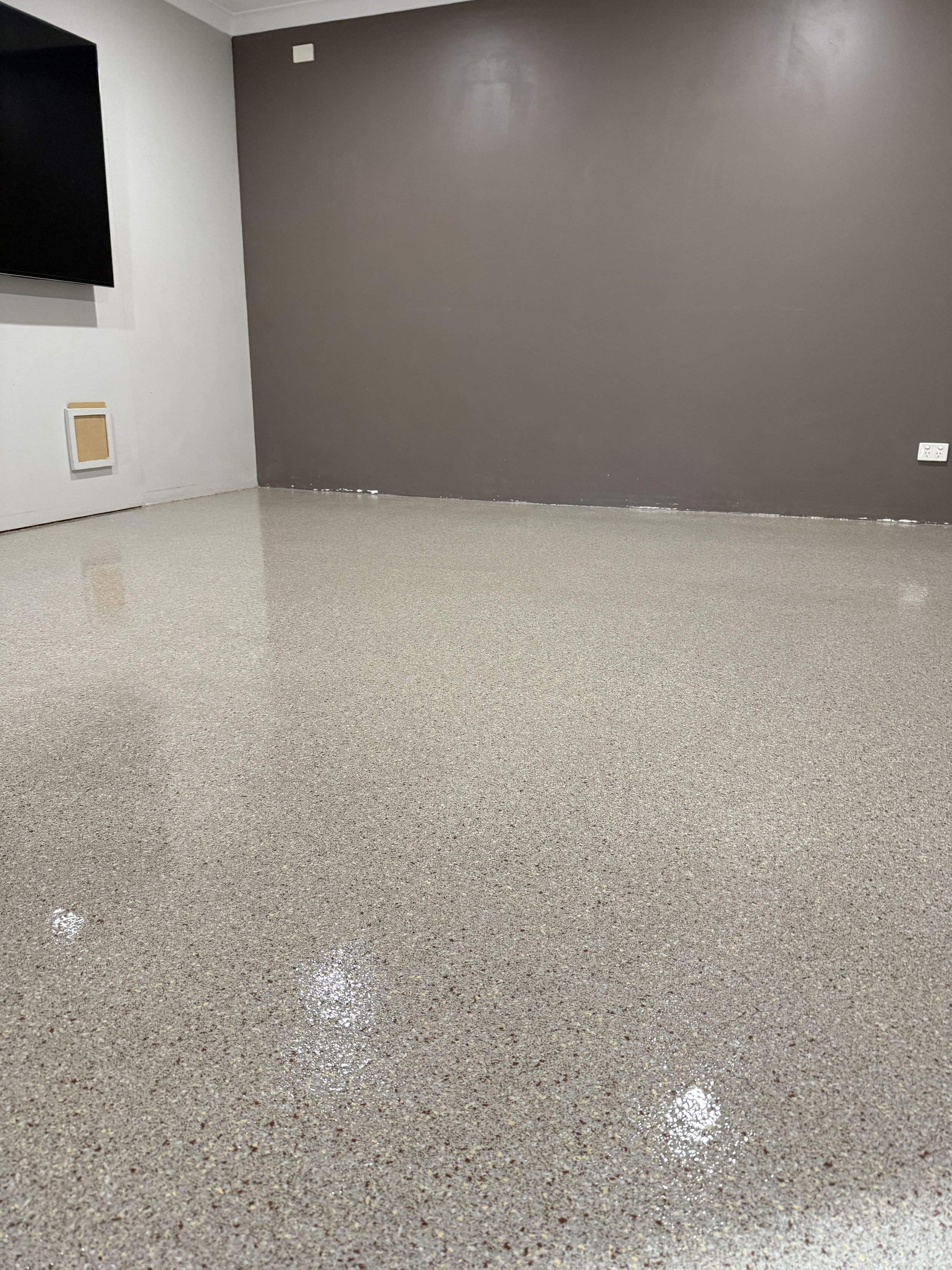 Polyaspartic floor Perth room with polished speckled beige floor, white and dark gray walls, a mounted television, and electrical outlets.