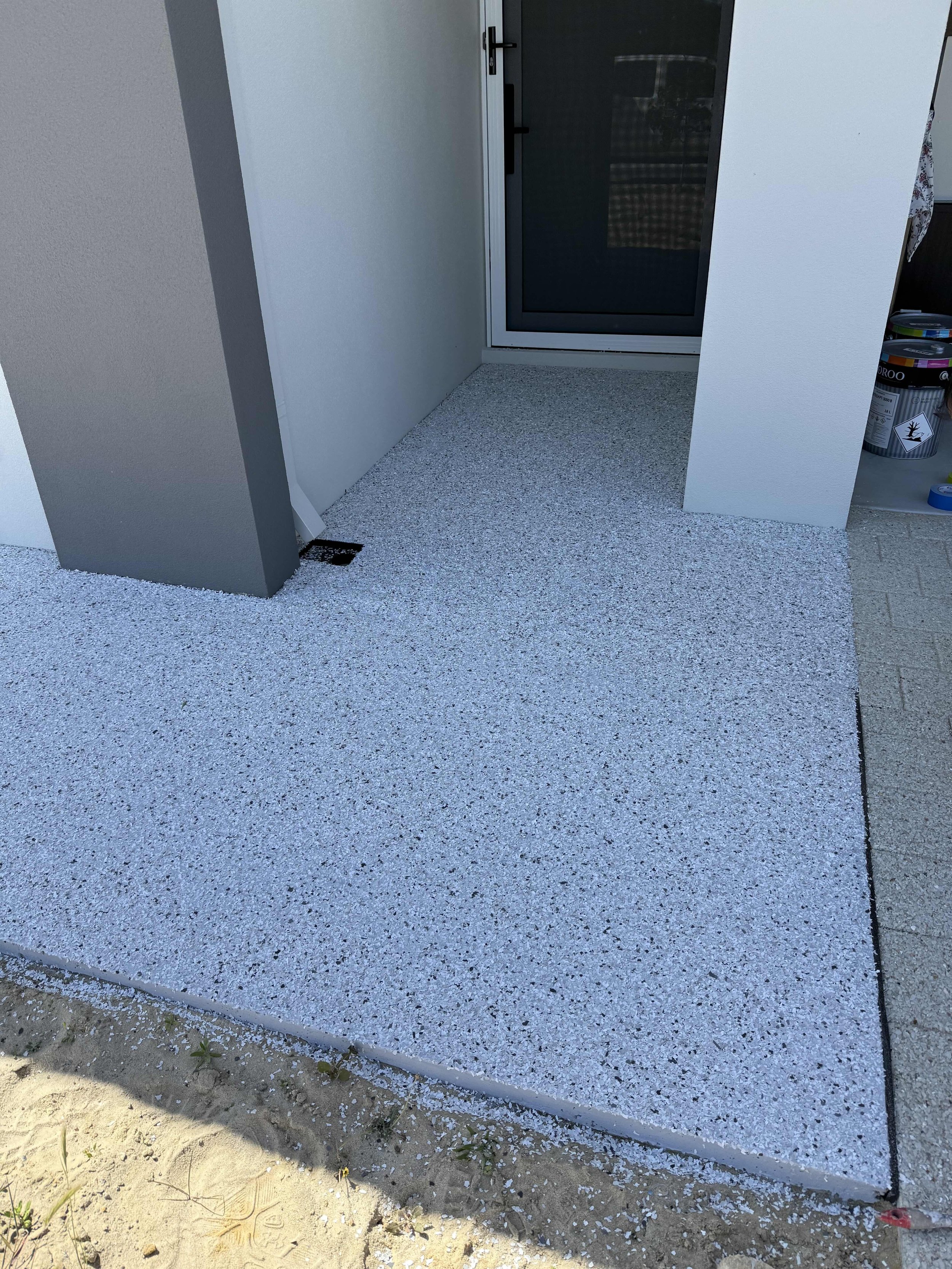 Newly installed grey and white epoxy flake with a sliding glass door, adjacent to a white and grey wall, and some garden bed dirt in the foreground.