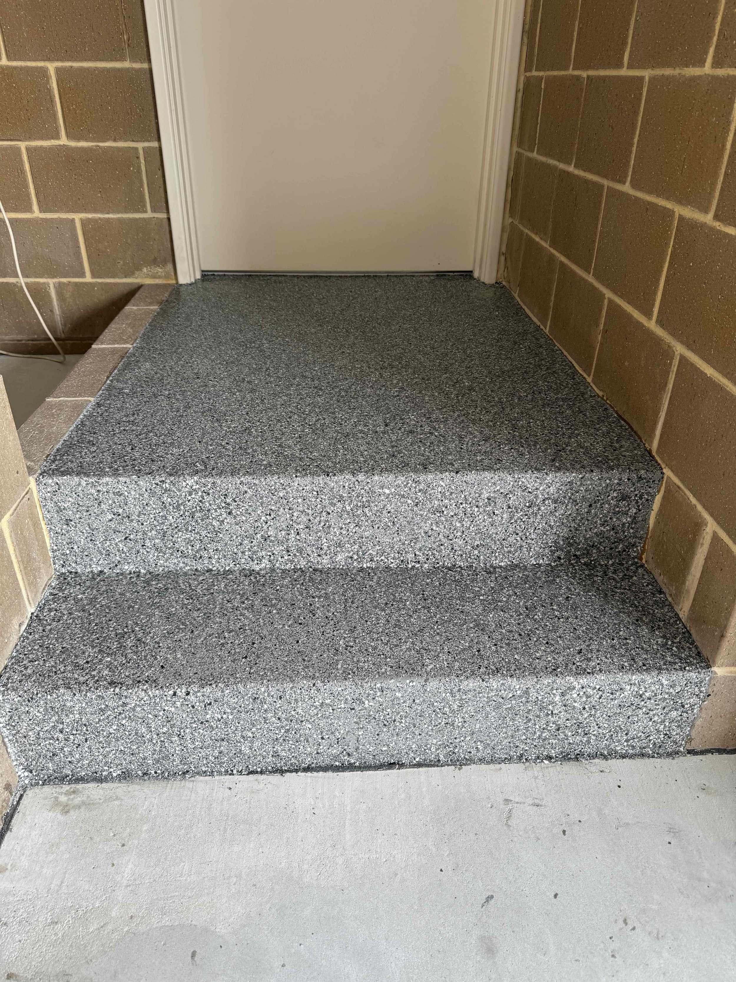 Concrete step with epoxy flakes with three levels leading up to a white door, surrounded by brick walls.