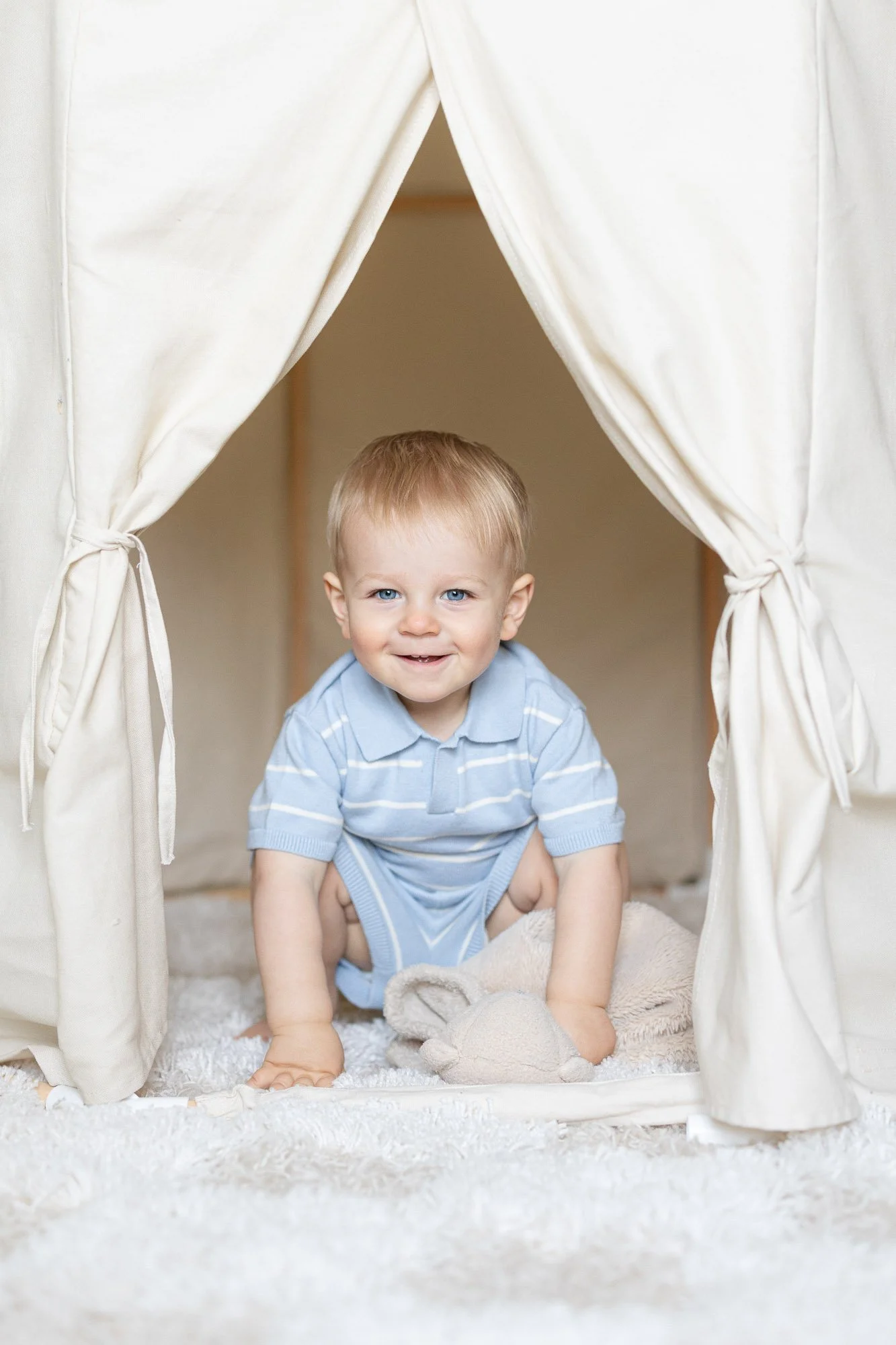 toddler boy in blue knit striped outfit smiling from inside a white play tent