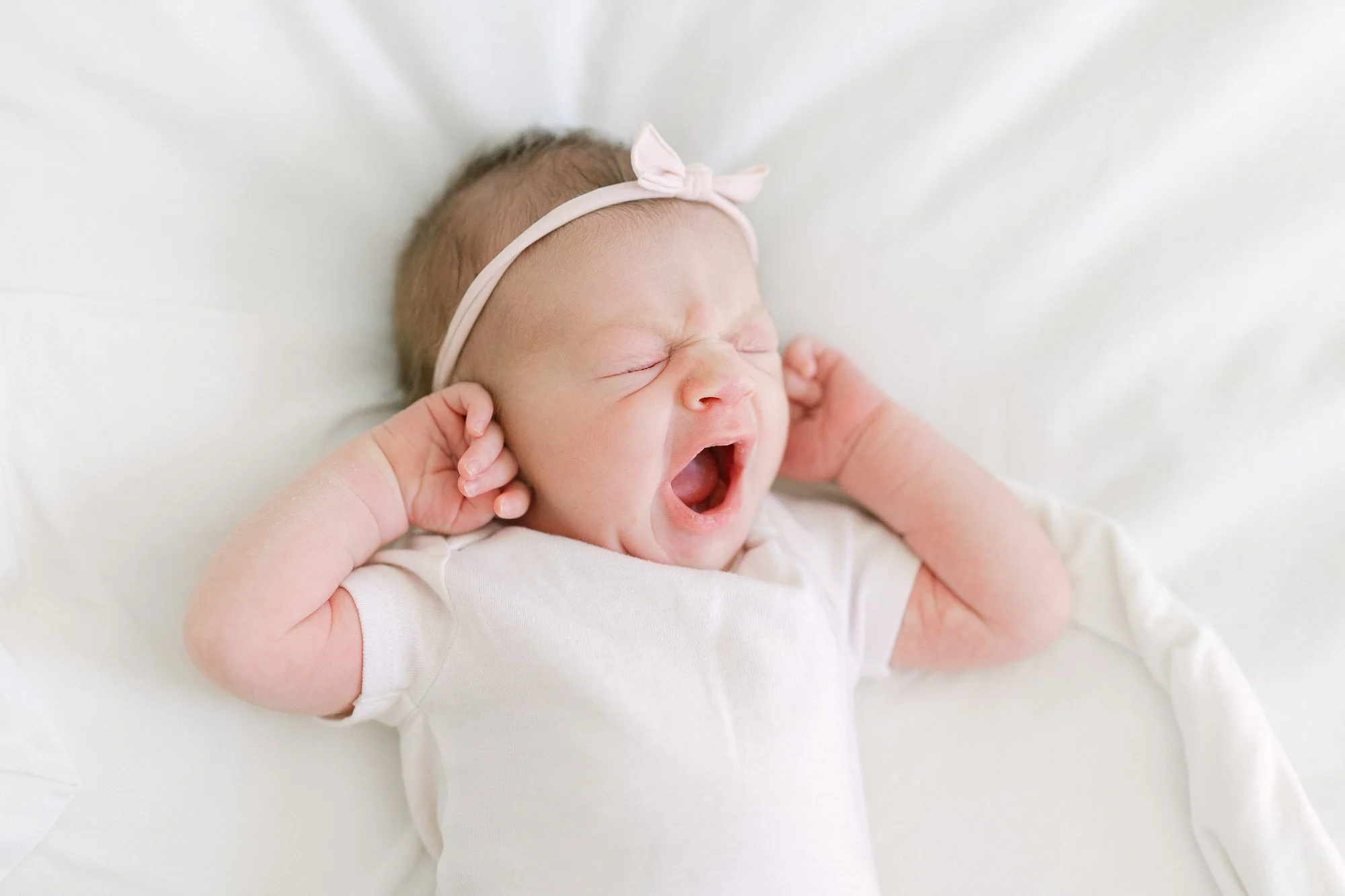 baby in white onesie and minimalistic bow yawning while lying on a bed with white linens during a home newborn session in Arlington Heights
