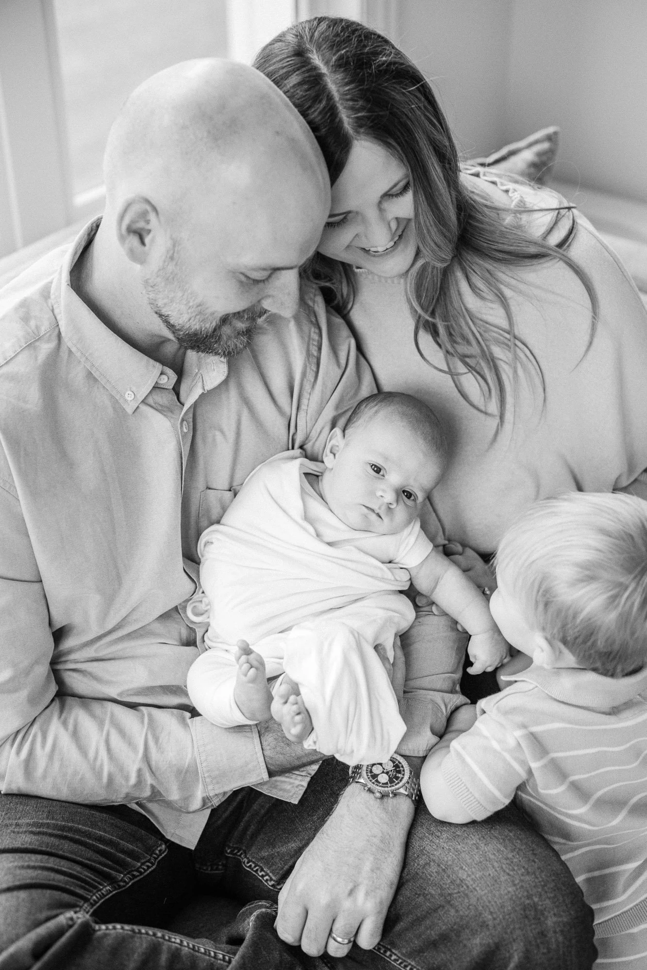 black and white edit of a mom and dad holding their newborn son while their toddler soon leans in to peek at him during a lifestyle newborn session in Hinsdale