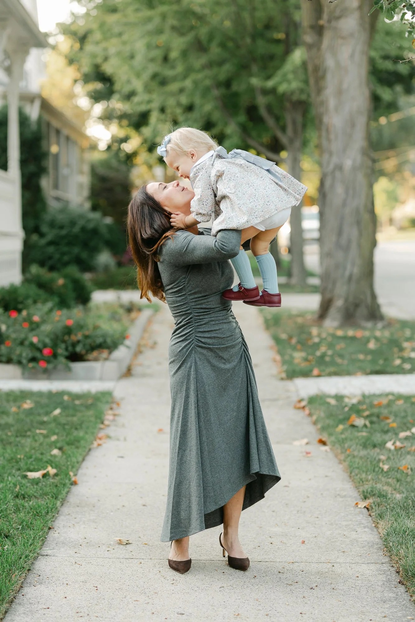 mom in a gray dress and high heels lifting her toddler daughter up to give her a kiss during an outdoor family session in downtown Arlington Heights