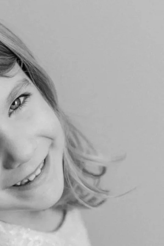 Close-up of a smiling young girl's face with light-colored hair, with part of her eye and hair visible in the image, in black and white.