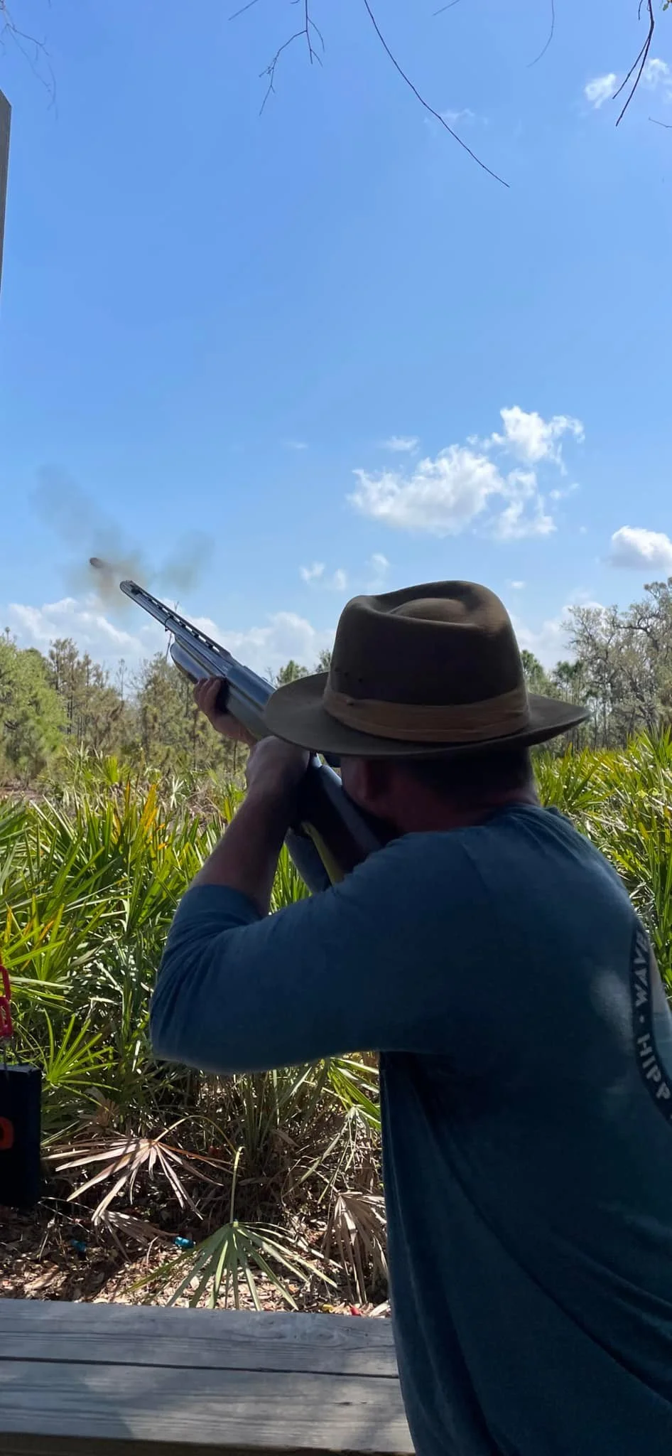 Man aiming a shotgun outdoors in a field with greenery and blue sky.
