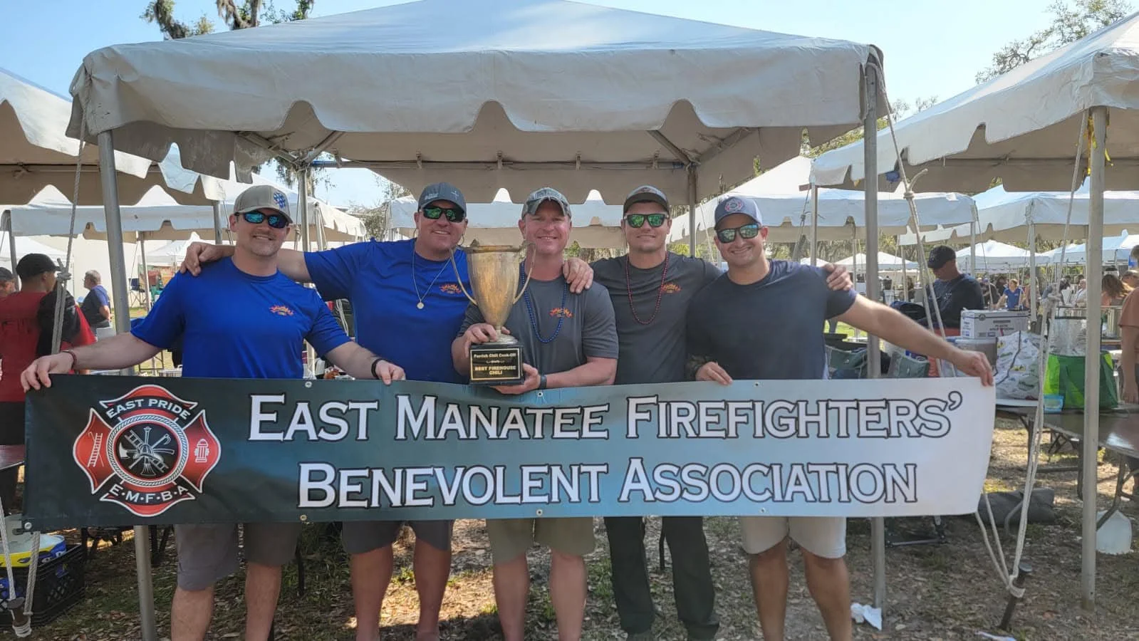 Group of five firefighters holding a trophy behind a banner that reads 'East Manatee Firefighters' Benevolent Association' at an outdoor event with tents and people in the background.