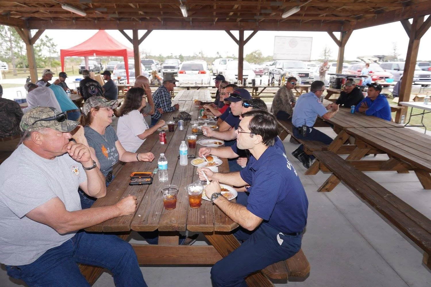 People sitting at a long wooden picnic table under a pavilion, eating and drinking during a gathering, with cars visible in the background.