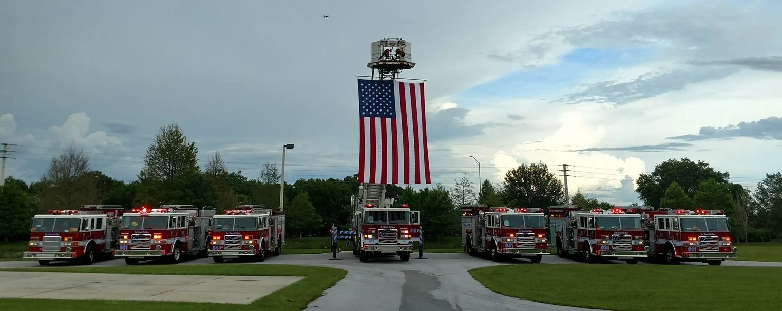 Seven fire trucks parked outdoors with a large American flag hanging from an aerial ladder in the center, against a cloudy sky
