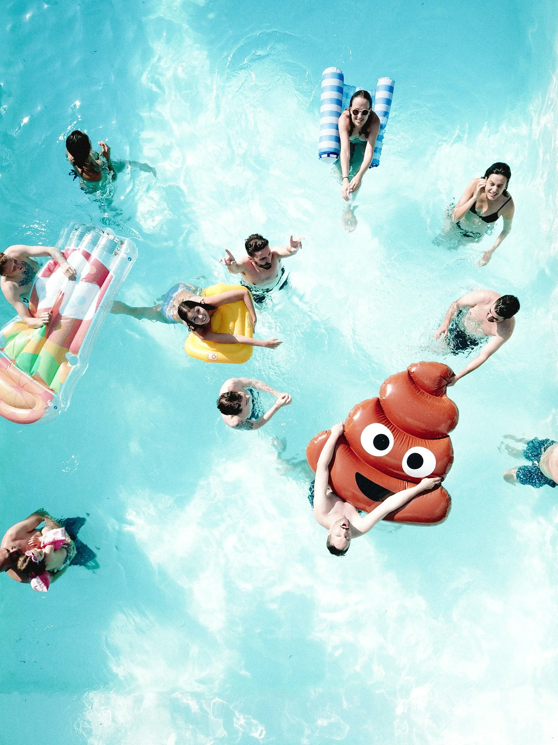 People enjoying a pool party with floats, including a person dressed as a smiling poop emoji, under bright sunlight.