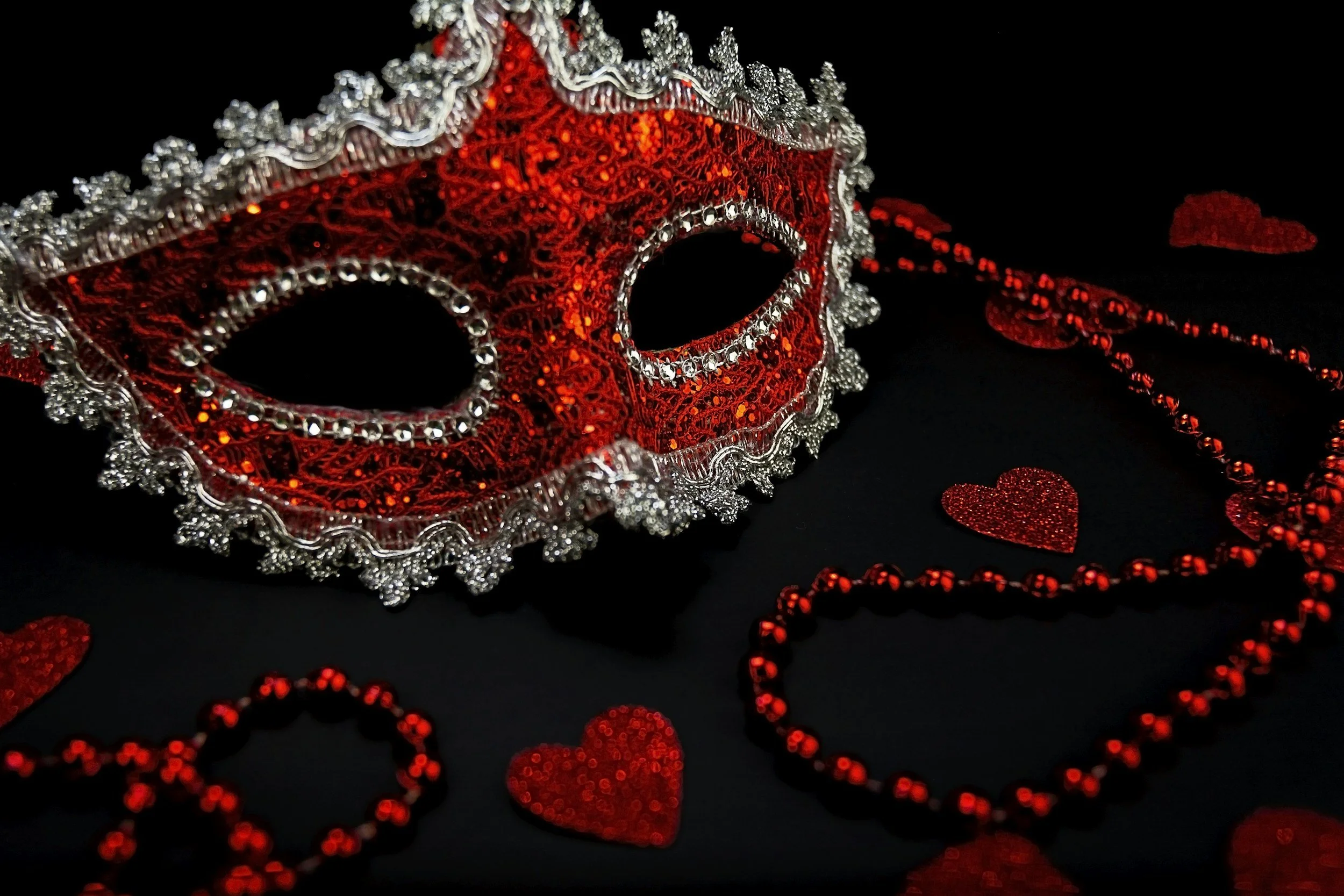 A red and silver decorative mask with intricate details, surrounded by red glittery hearts and beaded necklaces on a black background.