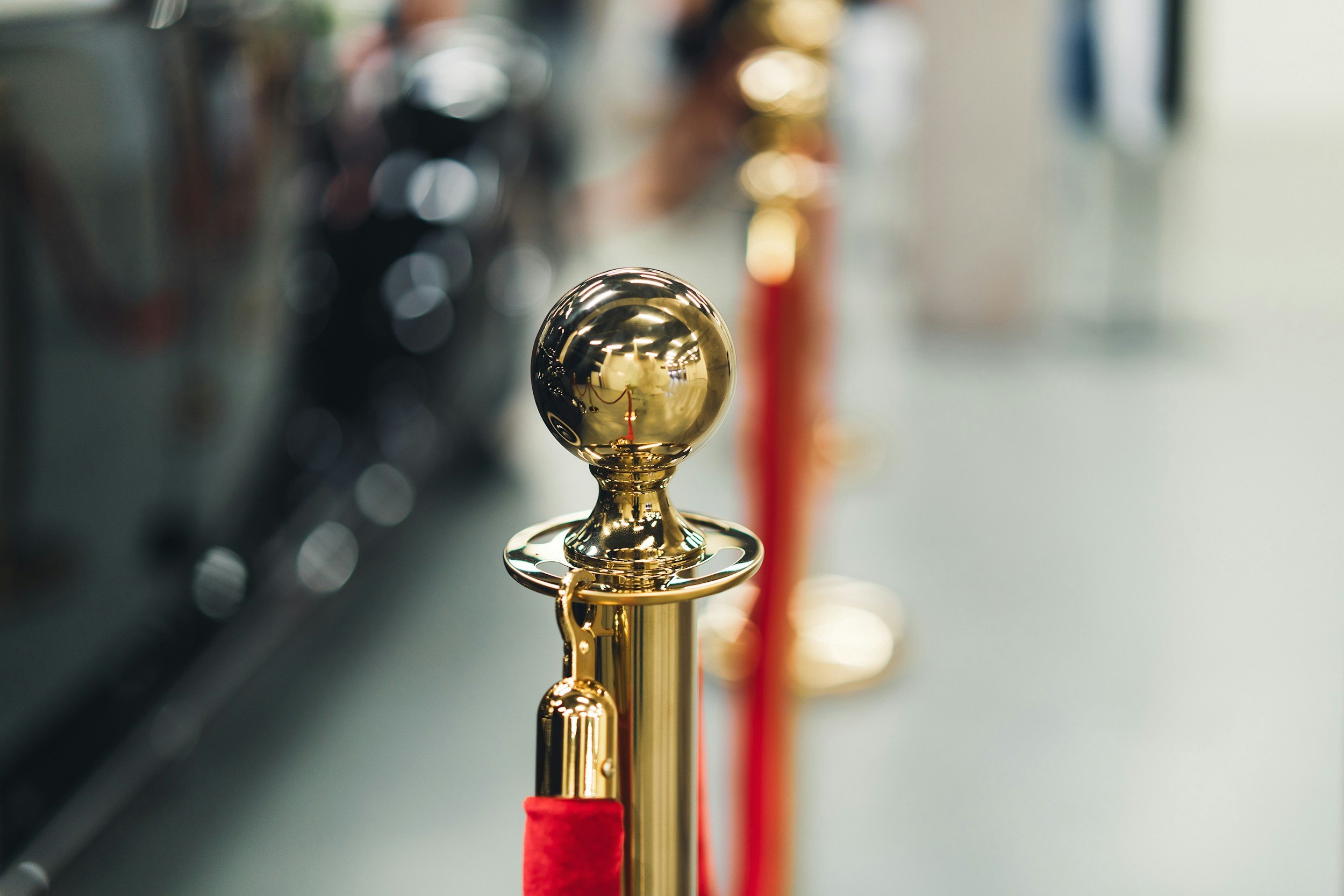 A shiny, golden metal post with a reflective spherical top and a red velvet rope attached, part of a queue barrier.