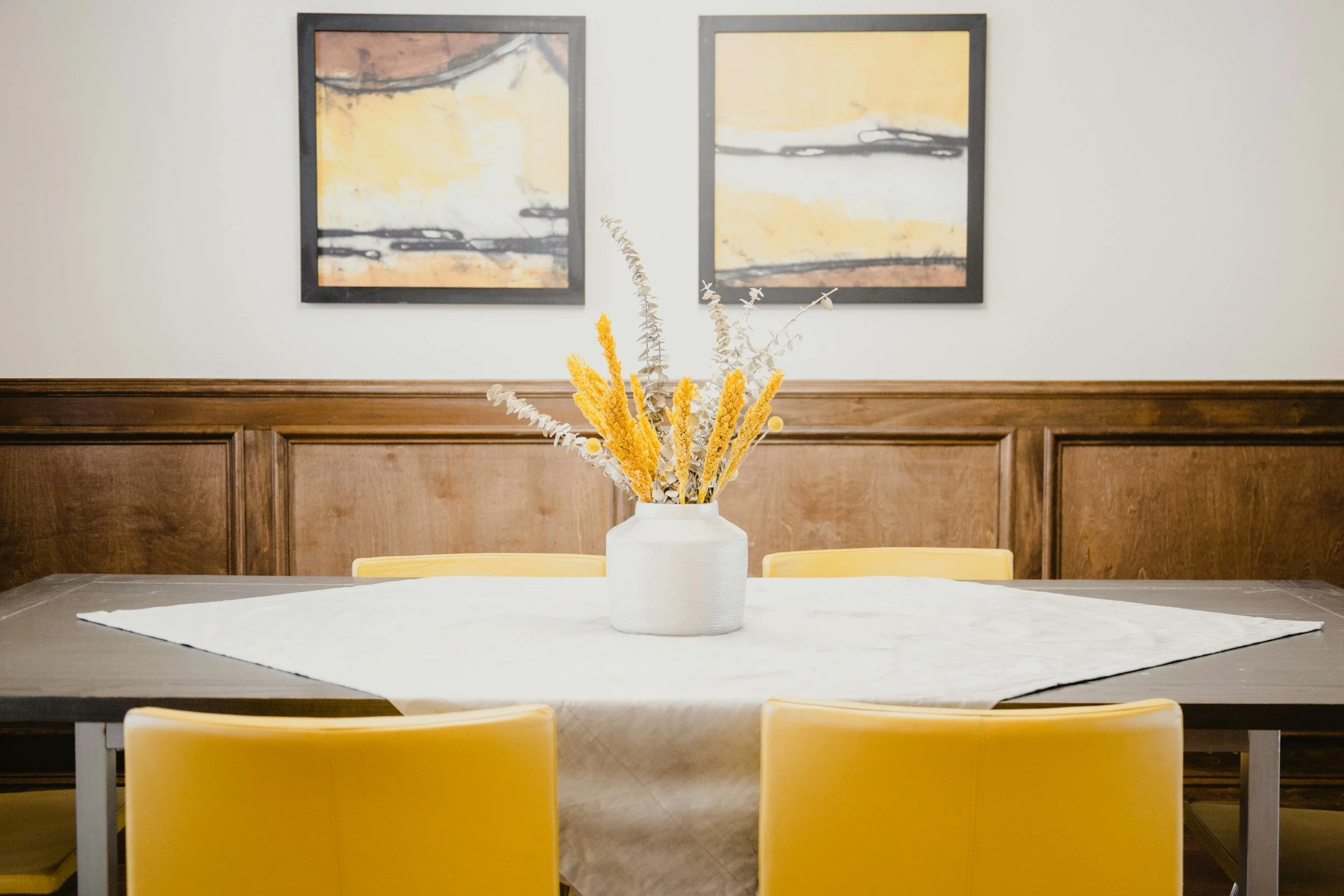 Dining room with a rectangular table, yellow chairs, a white vase with dried flowers as a centerpiece, a white tablecloth, and abstract artwork on the wall.